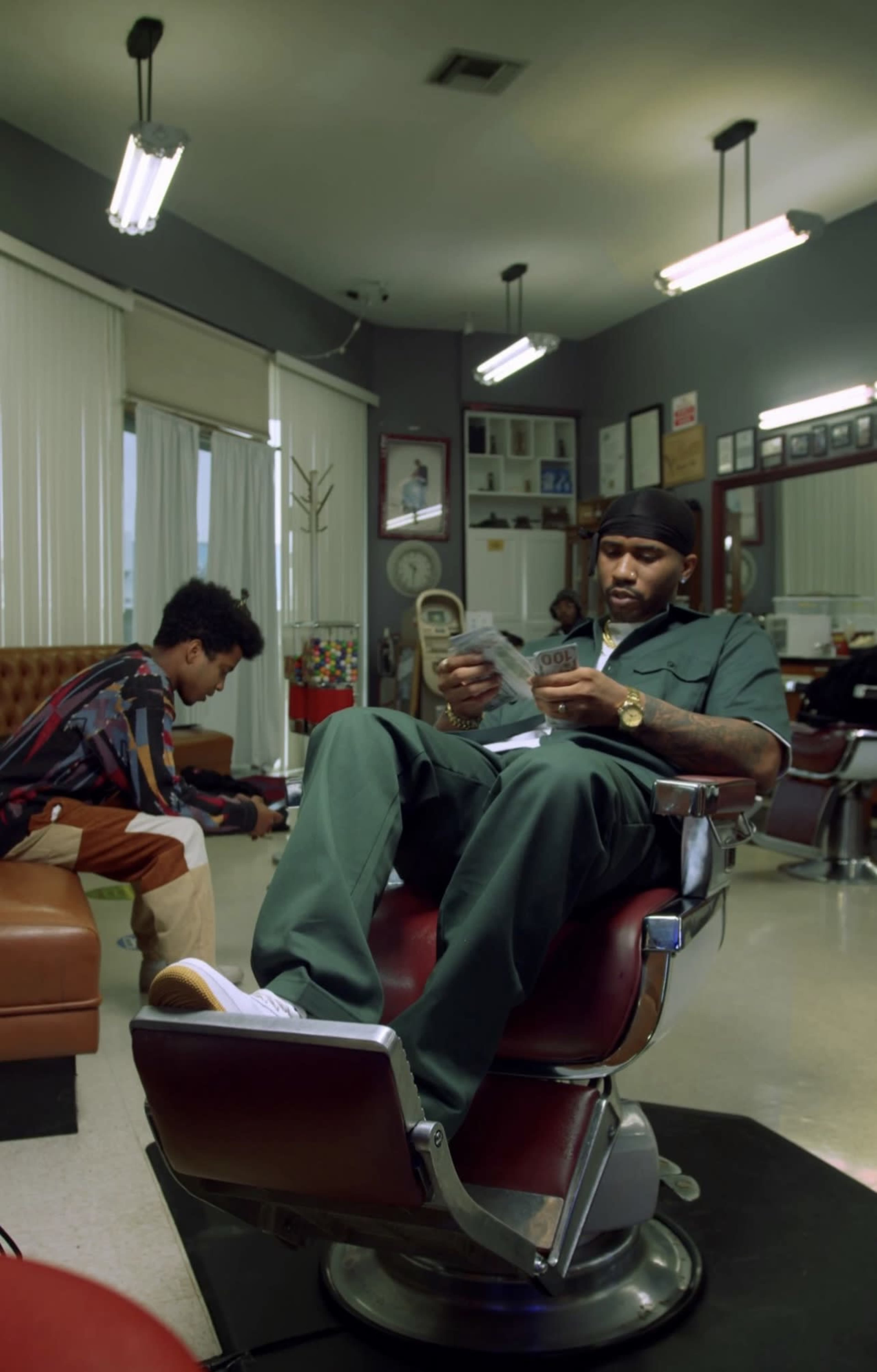 A man sits in a barber chair reading a magazine while a boy plays on the floor in a barbershop.