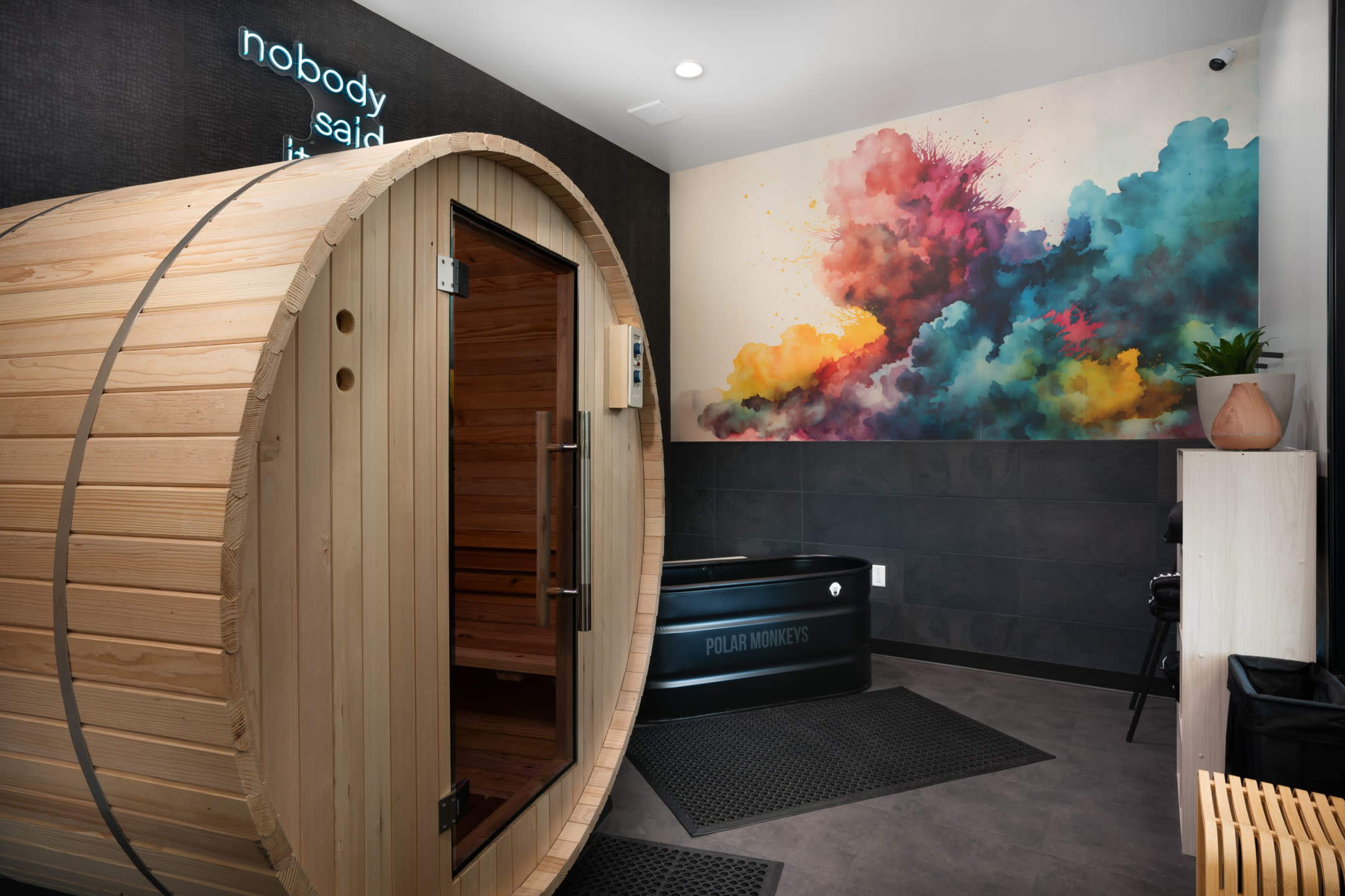 The image shows a modern wellness space featuring a wooden sauna barrel, a black cold plunge tub, and a colorful abstract mural on the wall.