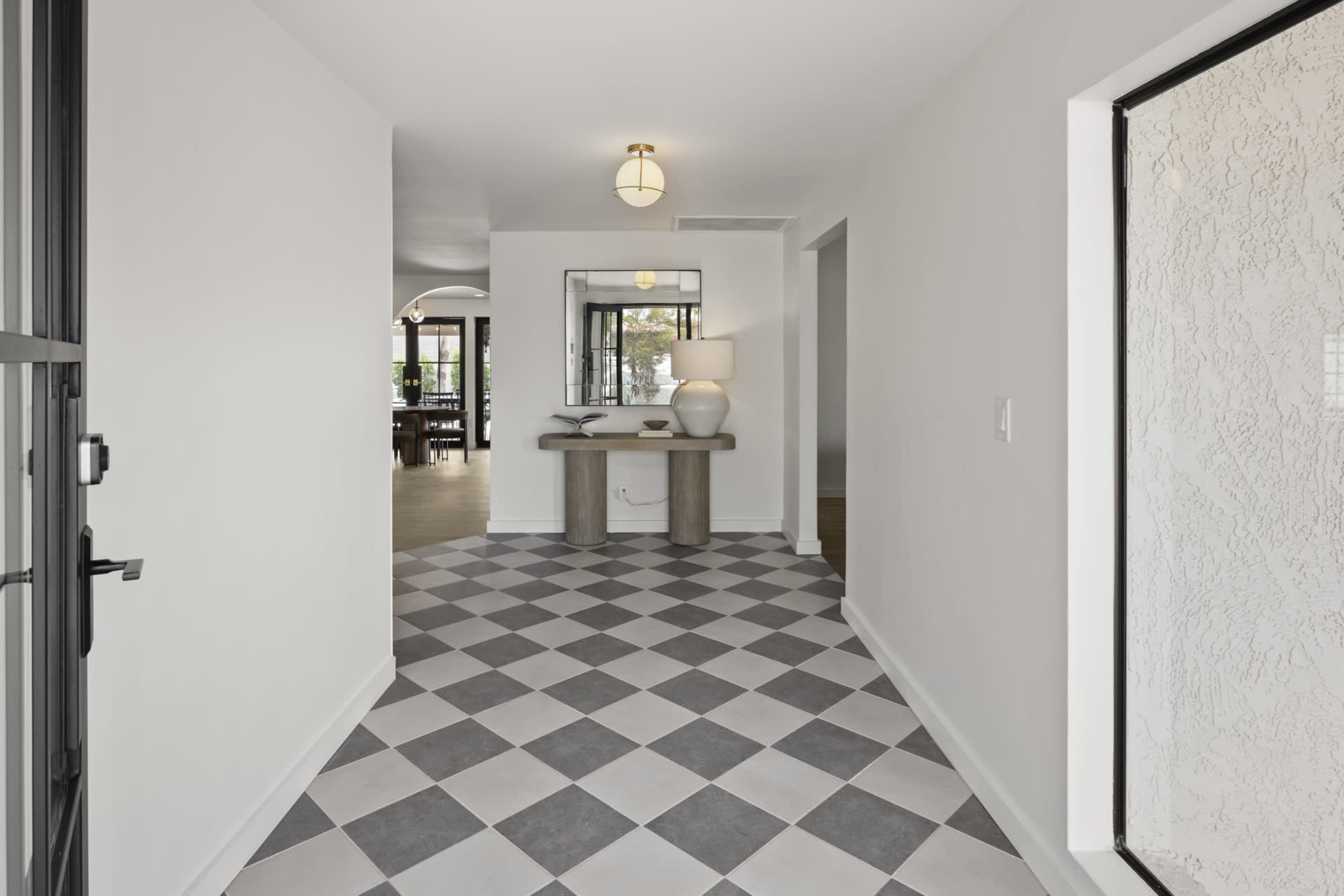 The image shows a spacious hallway with black and white checkered tile flooring, leading to an open area with a table and lamp against a white wall.