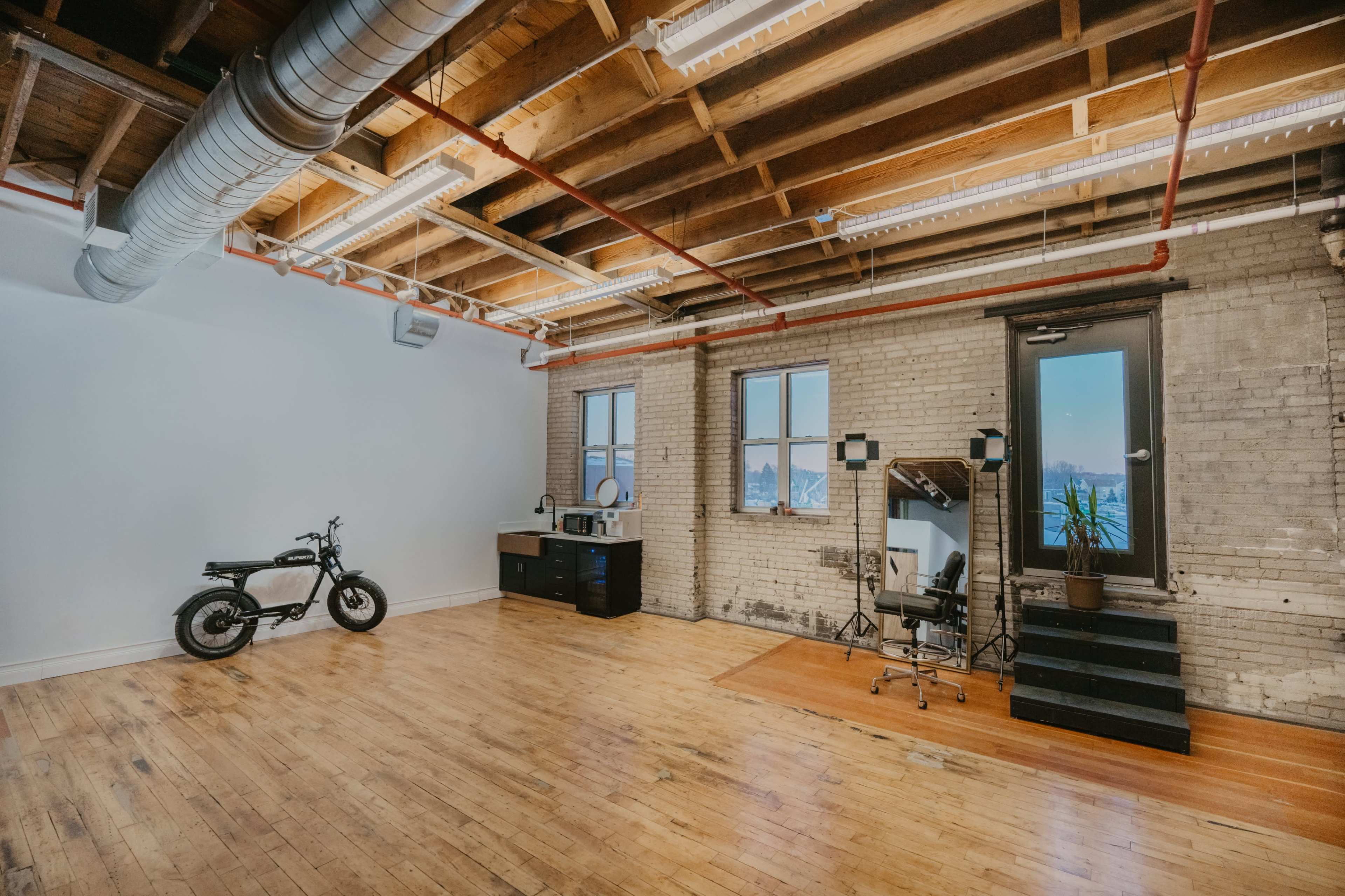 A spacious, industrial-style room features wooden flooring, exposed beams, and a brick wall, with a bicycle, a kitchenette, two chairs, and a staircase leading to a window.