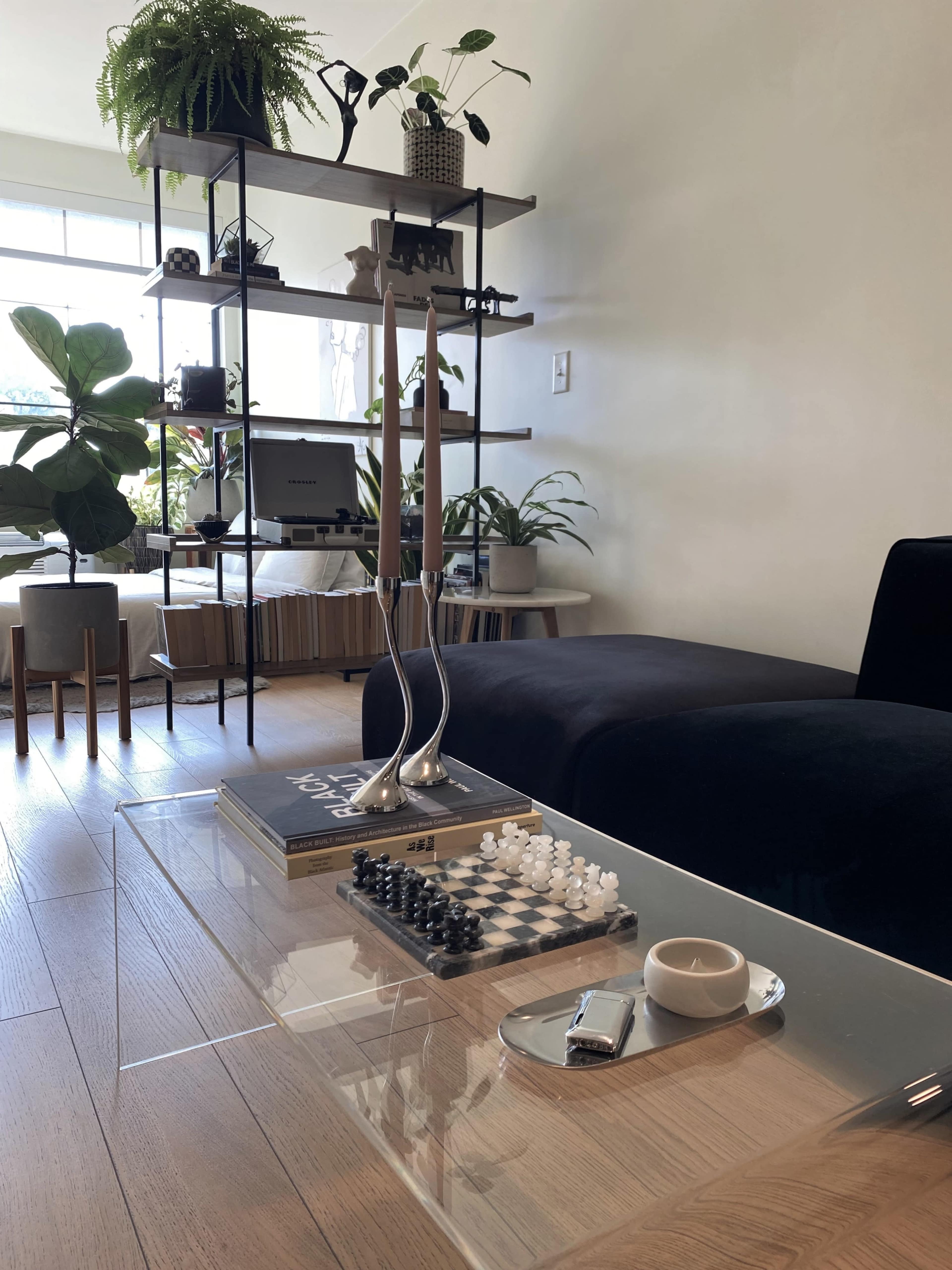 A modern living room features a glass coffee table with a chess set, a book, and a plate, surrounded by plants and stylish furniture.