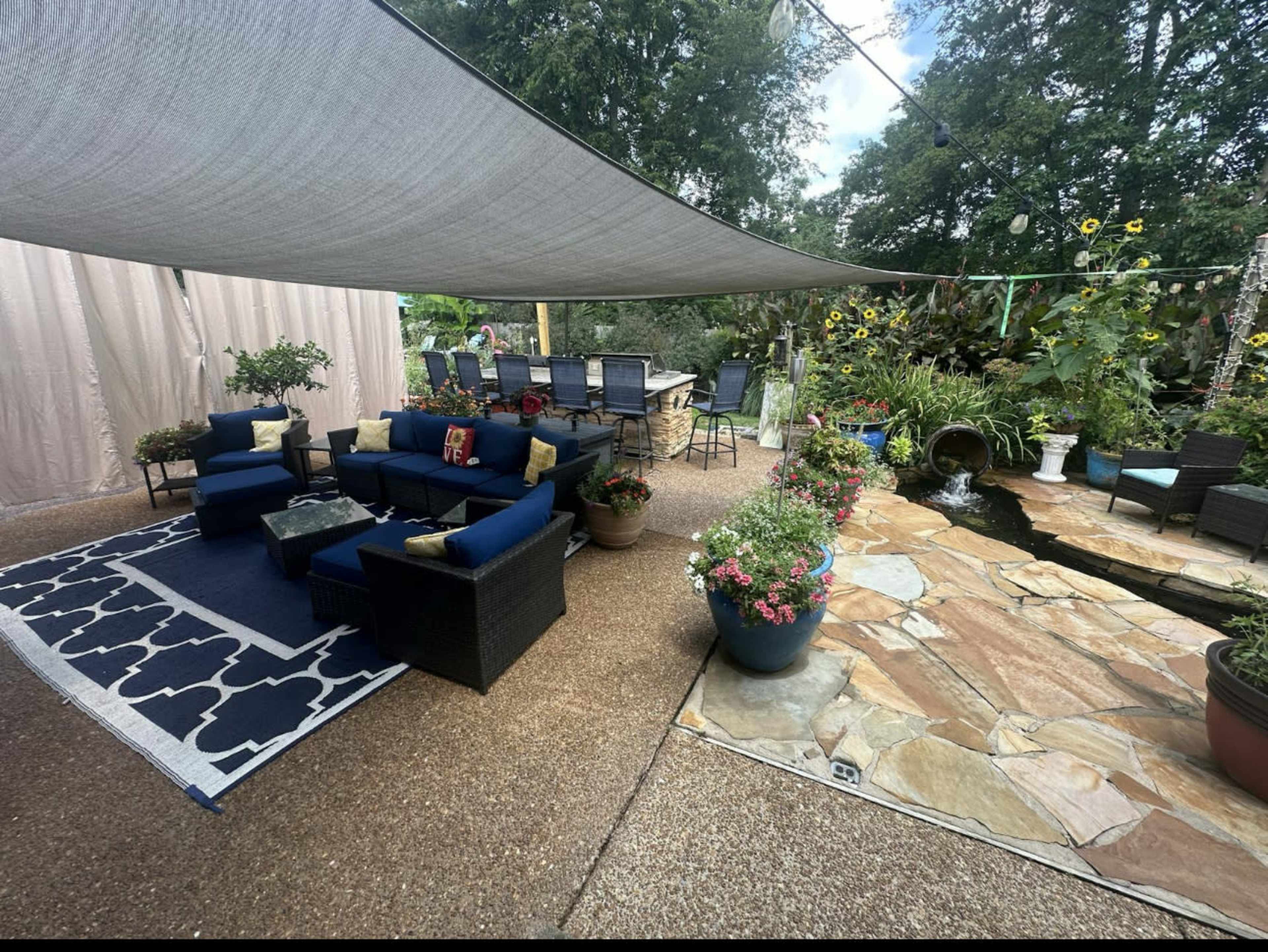 The image shows a landscaped outdoor seating area with blue upholstered furniture, a stone pathway, potted plants, and a shaded dining table nearby.
