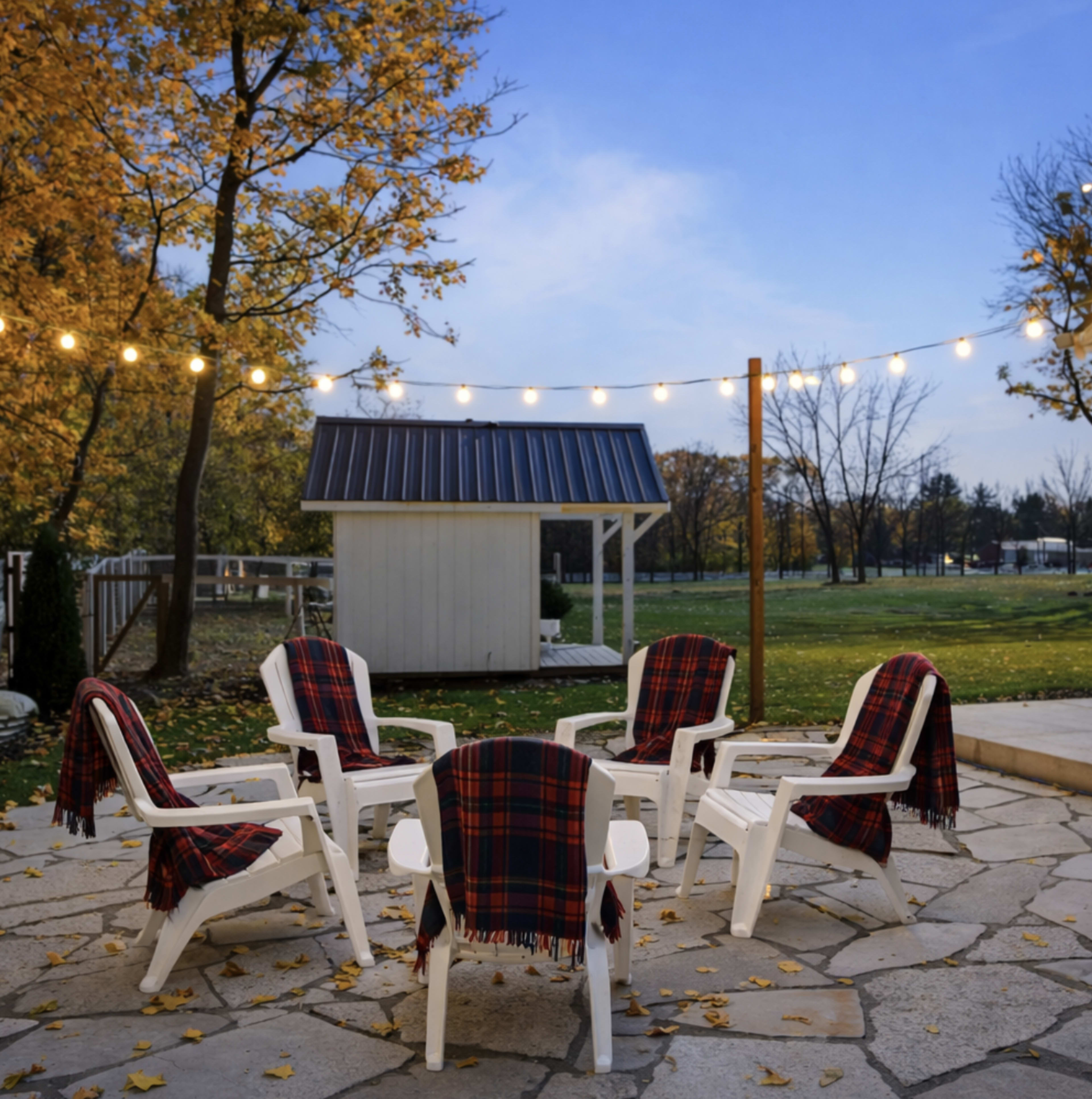 A circular arrangement of white chairs with plaid blankets draped over them is set on a stone patio, surrounded by trees with autumn foliage and string lights overhead.