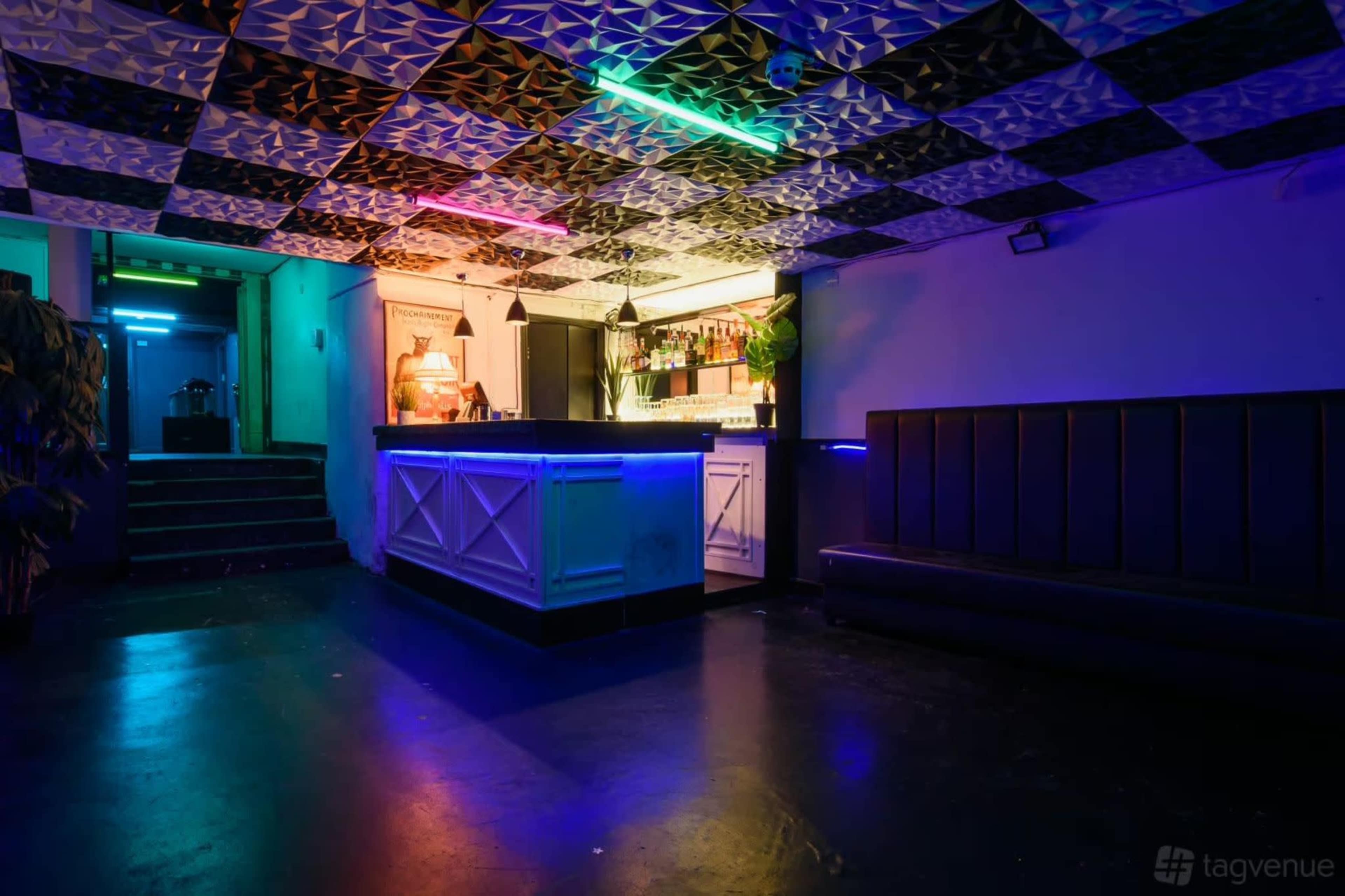 The image shows a dimly lit bar area with a patterned ceiling, illuminated by colorful lights, and a sleek counter behind which a selection of bottles is displayed.