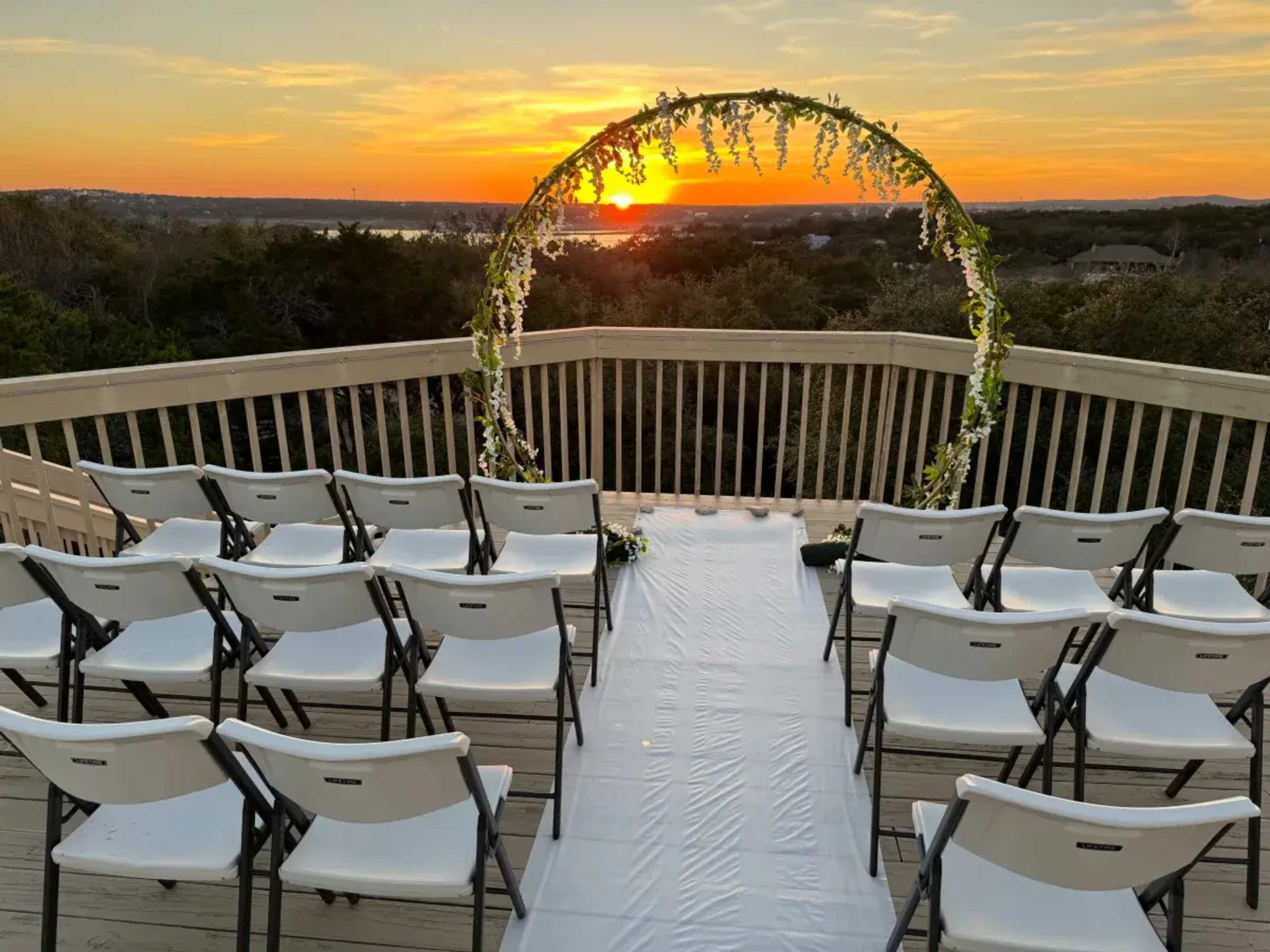 A wedding venue features white chairs set in a semicircle around a flower-adorned arch, overlooking a sunset skyline.