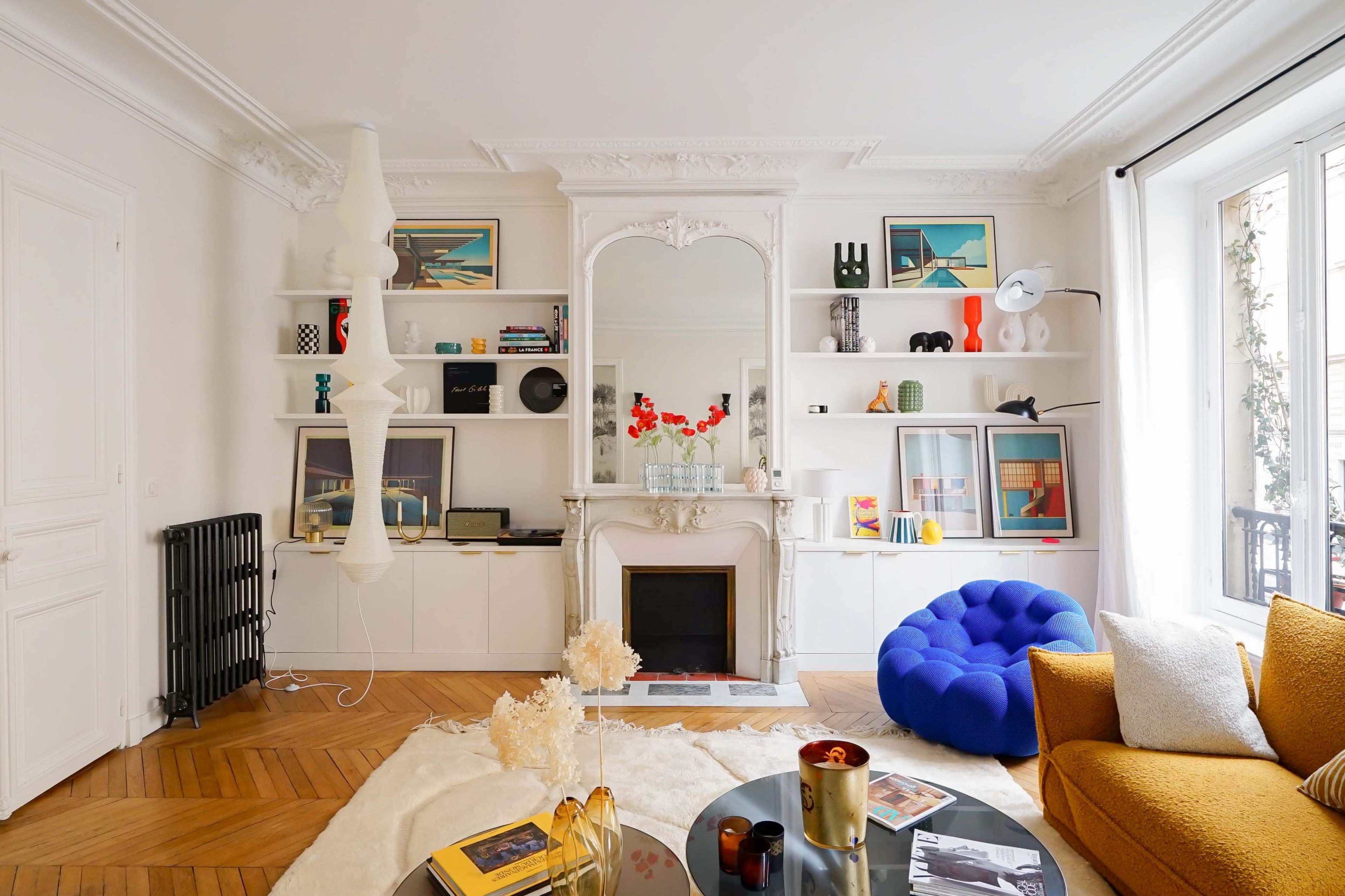 The image shows a neatly arranged living room featuring a central fireplace, built-in shelves with decor, a large blue pouf, and a mustard-colored sofa, all set against hardwood floors.