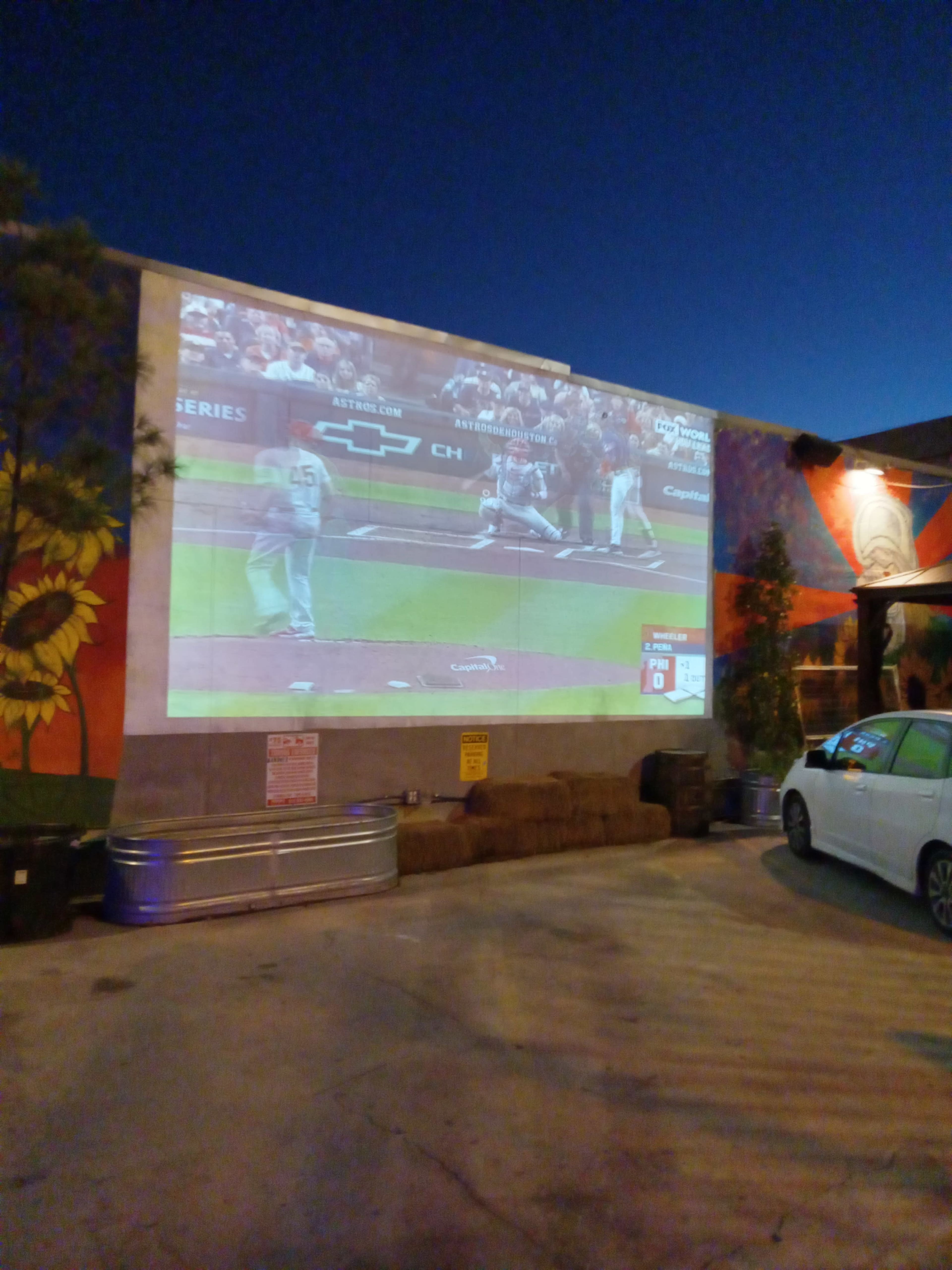 A baseball game is being projected onto a wall in an outdoor setting, with visible seating and artistic murals nearby.