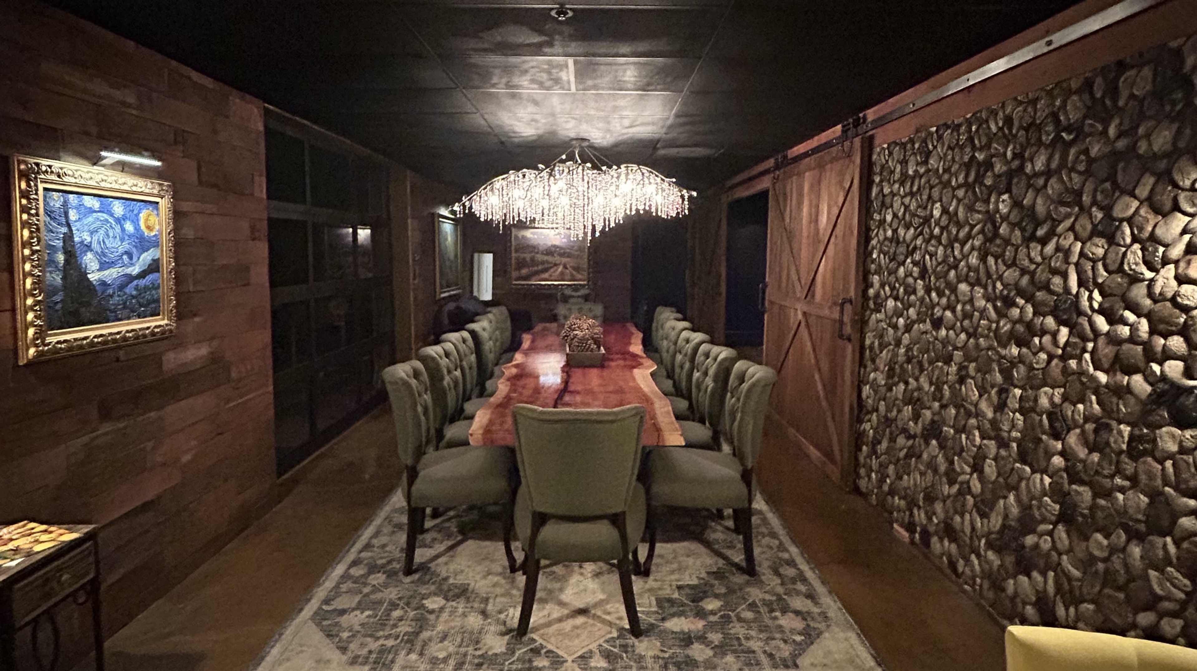 A long wooden table surrounded by green chairs is set in a dimly lit dining room with stone and wood walls.