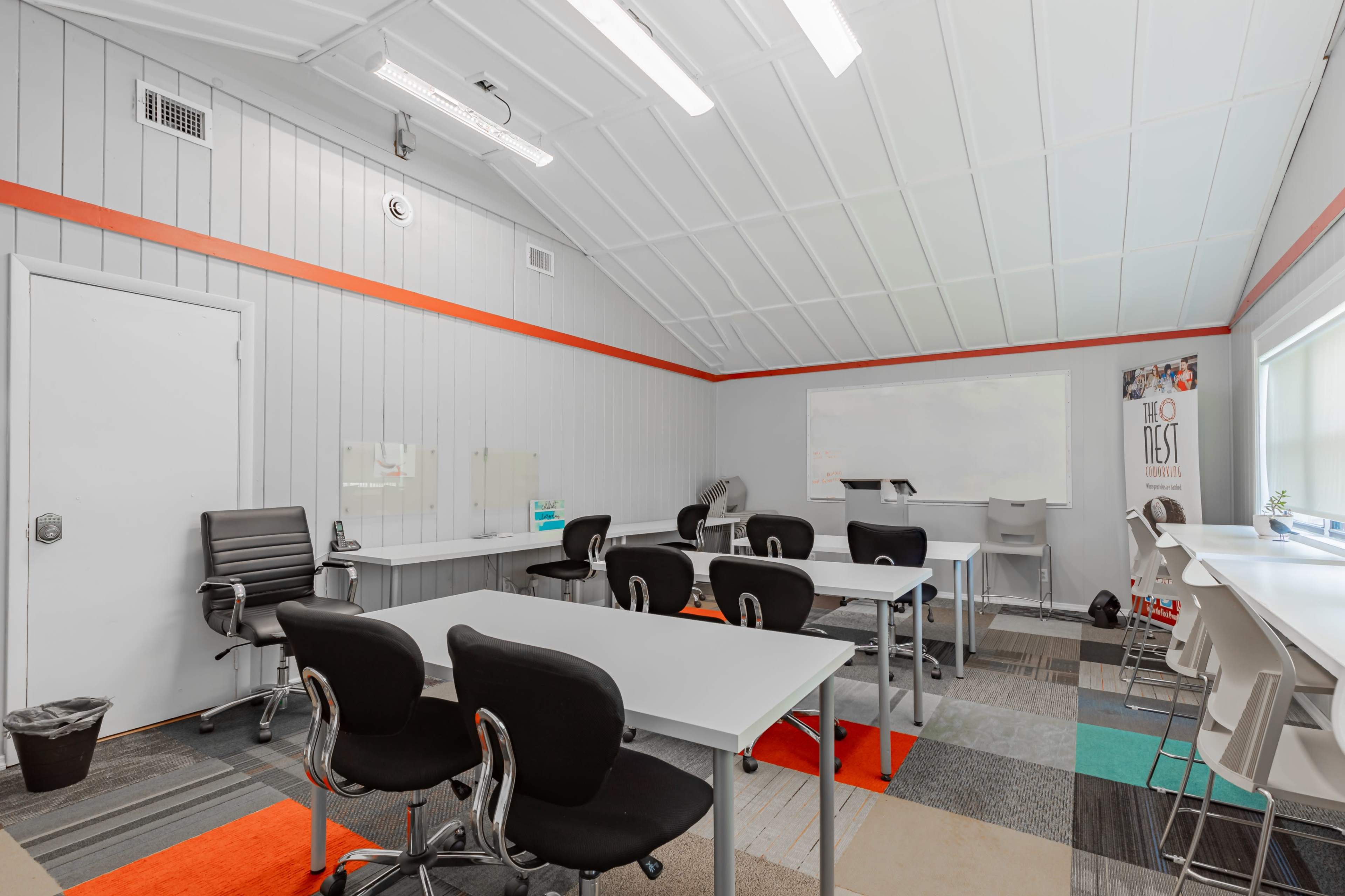 A well-lit classroom features several white tables and chairs arranged around a central area with colorful carpet tiles and a whiteboard on the wall.