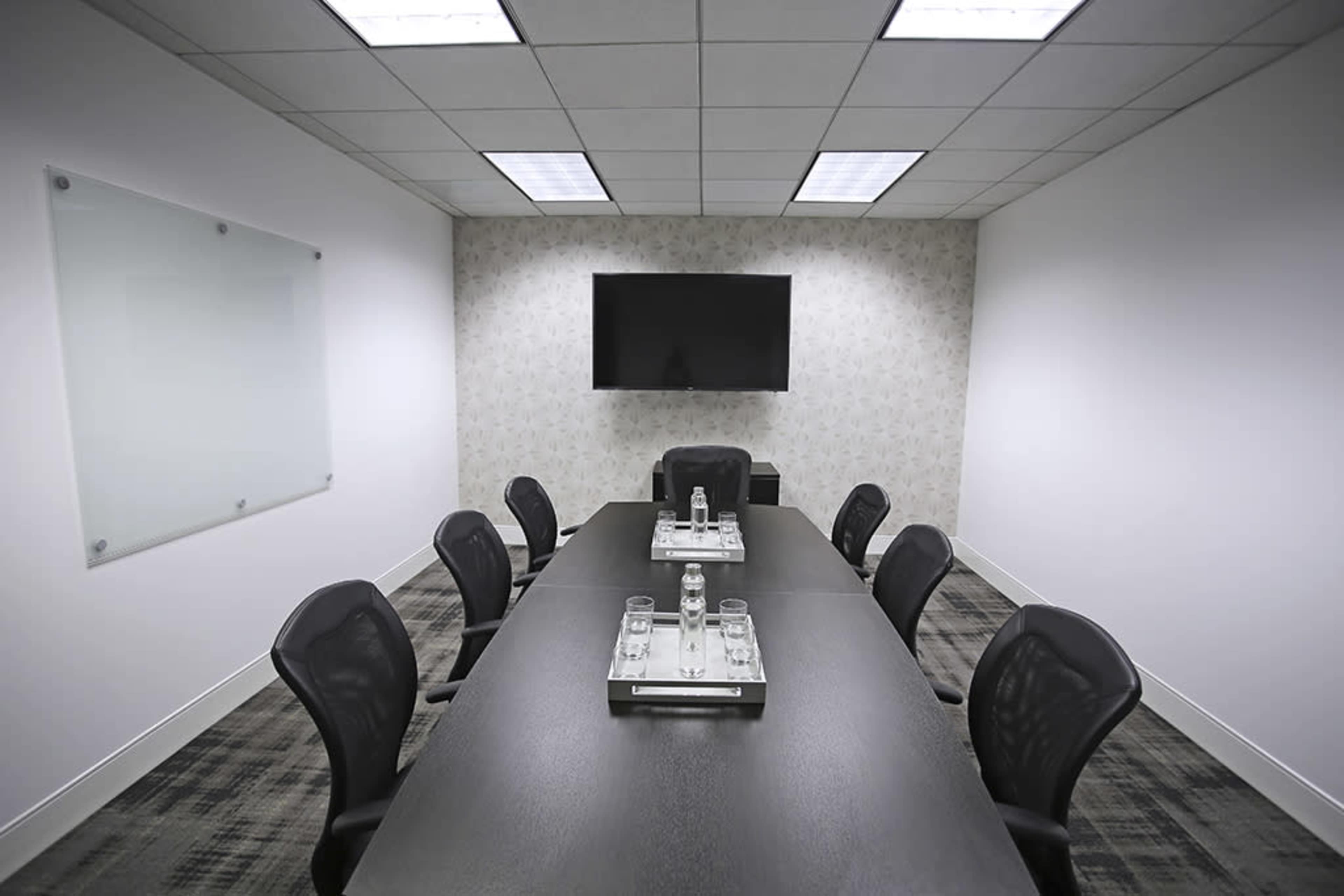 A conference room features a long rectangular table surrounded by black chairs, with a television mounted on the wall and two glass water bottles placed on a tray in the center.