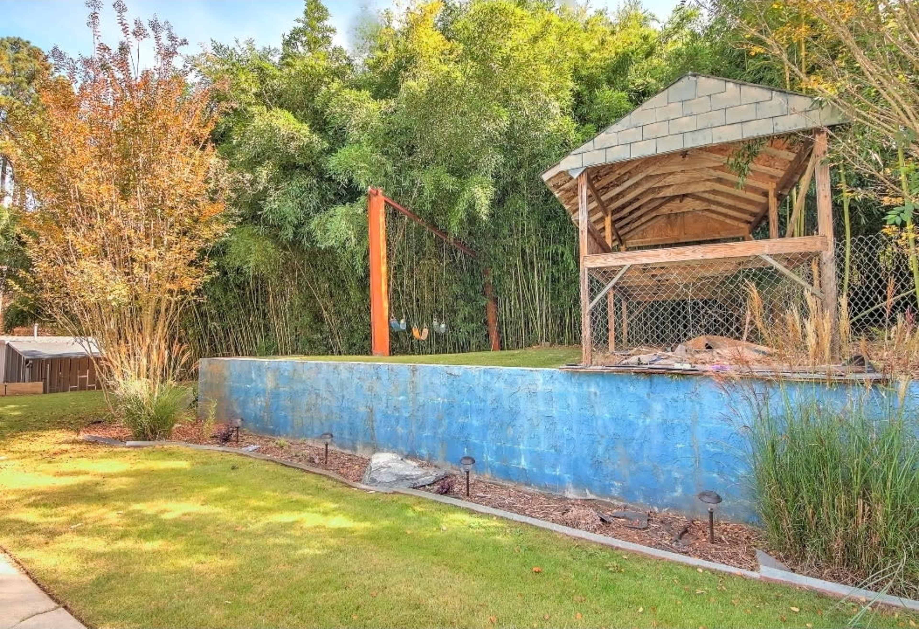 The image shows a grassy area with a blue stone wall, a dilapidated wooden structure, and bamboo plants in the background.