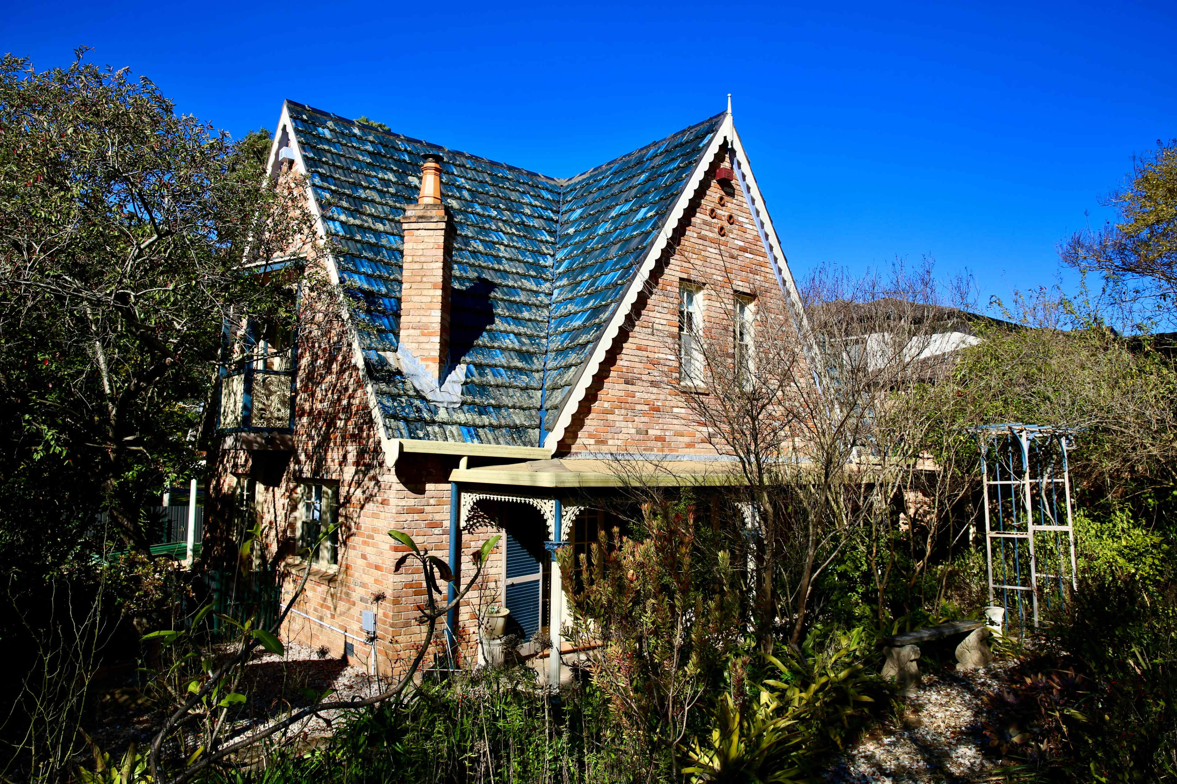 A brick house with a triangular roof, surrounded by trees and plants, is set against a clear blue sky.
