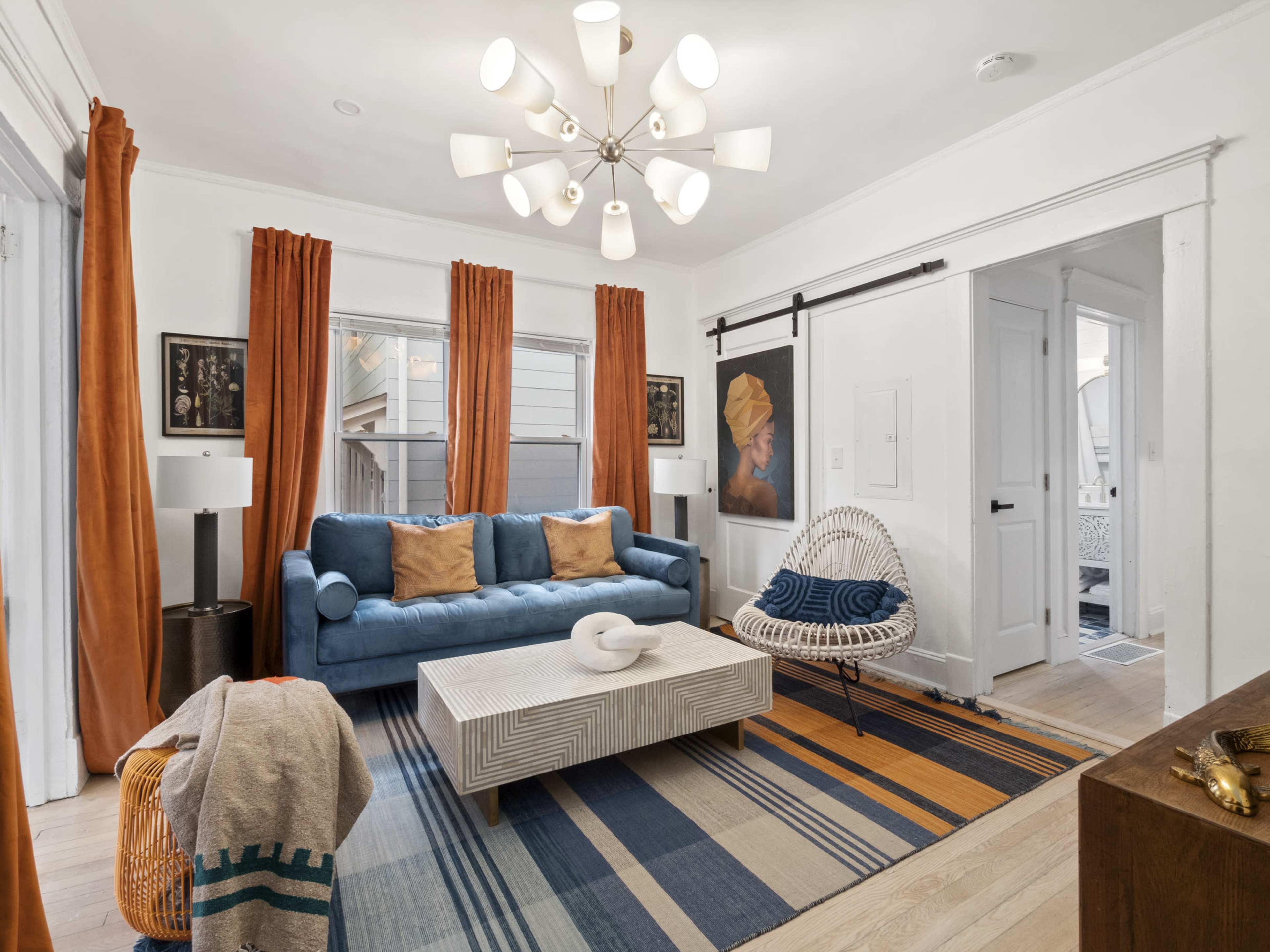A modern living room features a blue sofa, a patterned area rug, and large windows with orange curtains.