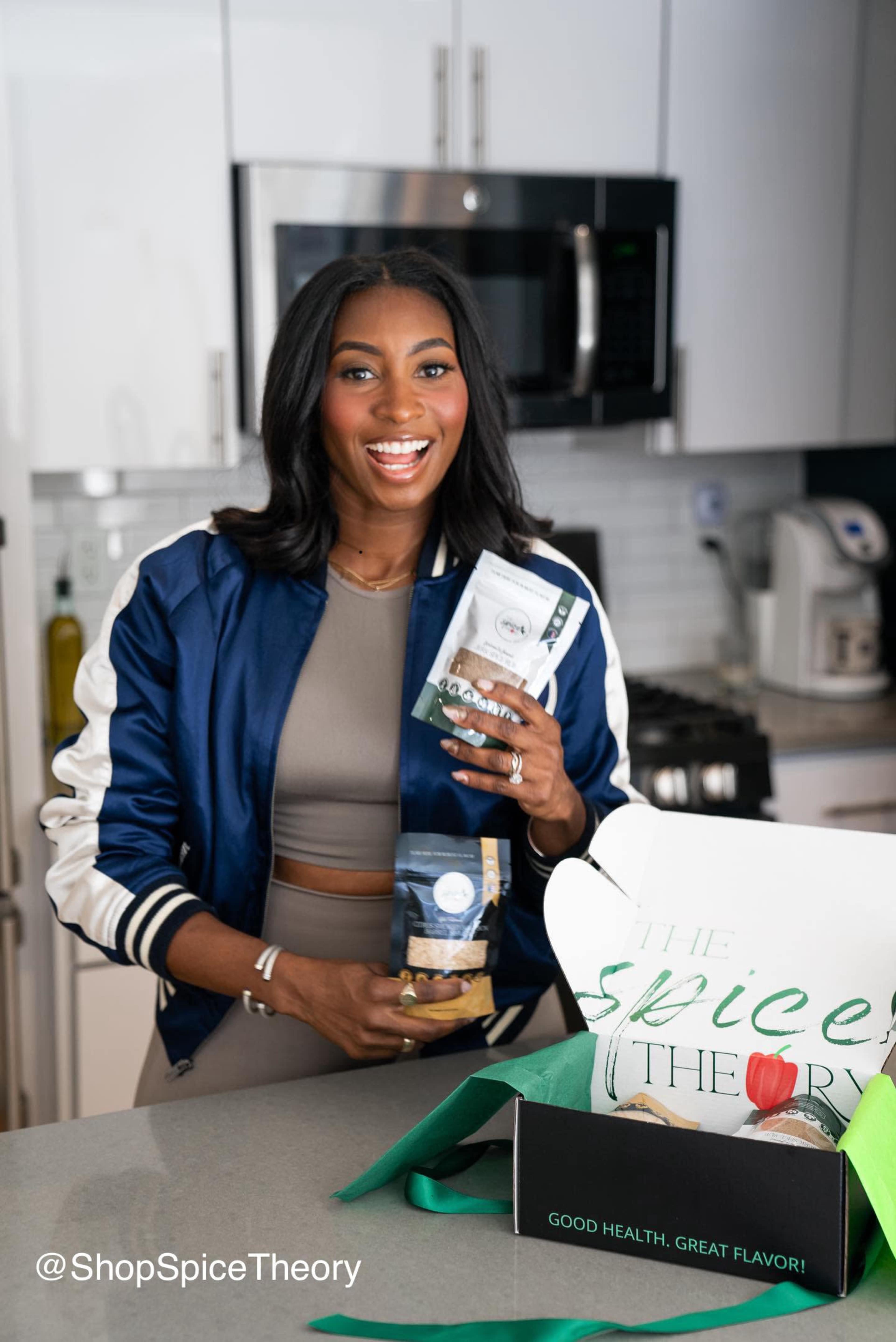 A woman in a blue jacket stands in a kitchen, holding spice packages in one hand and a box labeled "The Spice Theory" on the counter.