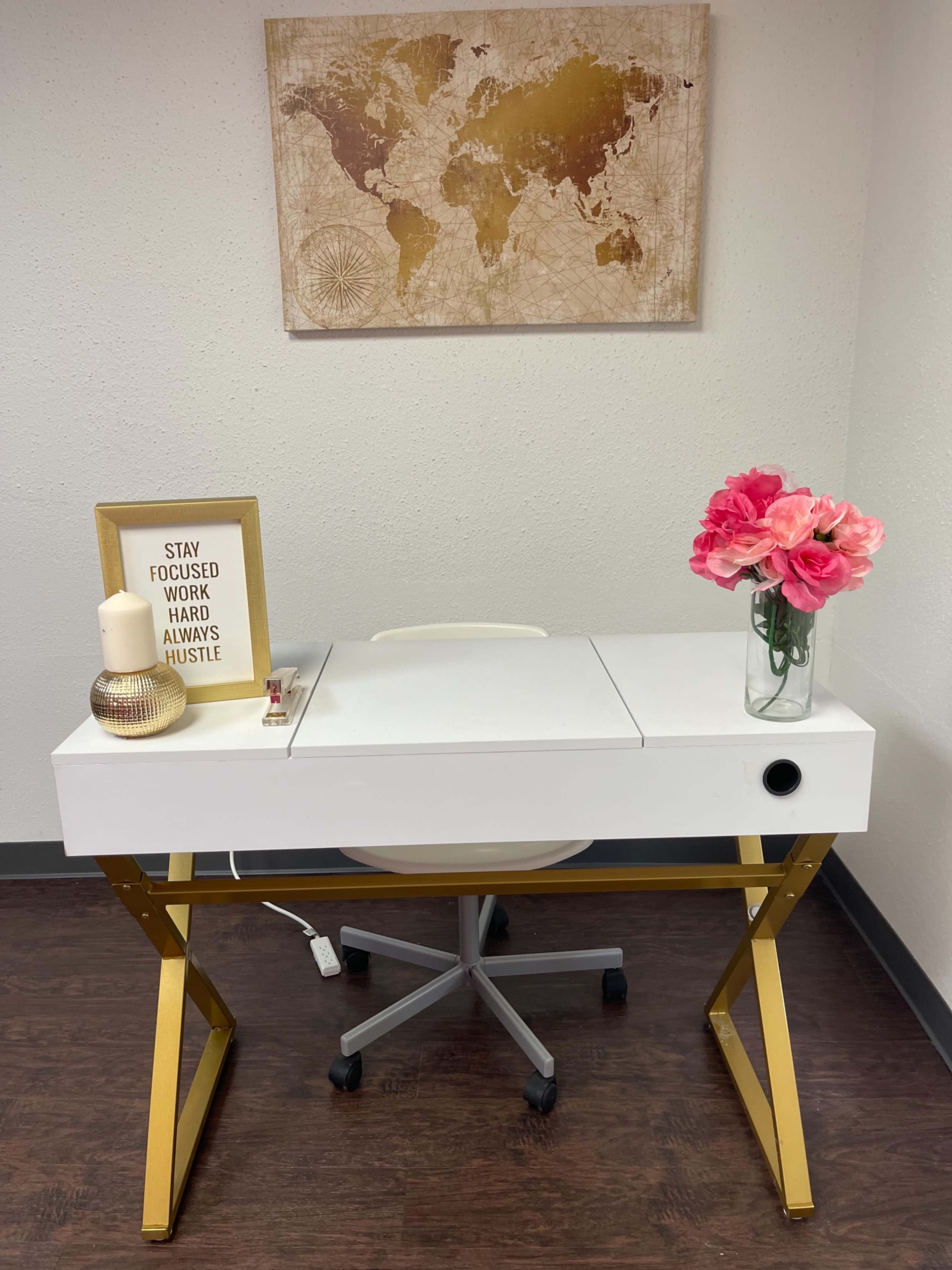 A white desk with a gold frame, accompanied by a chair, a framed motivational quote, a flower vase, and a candle, is set against a wall with a world map.