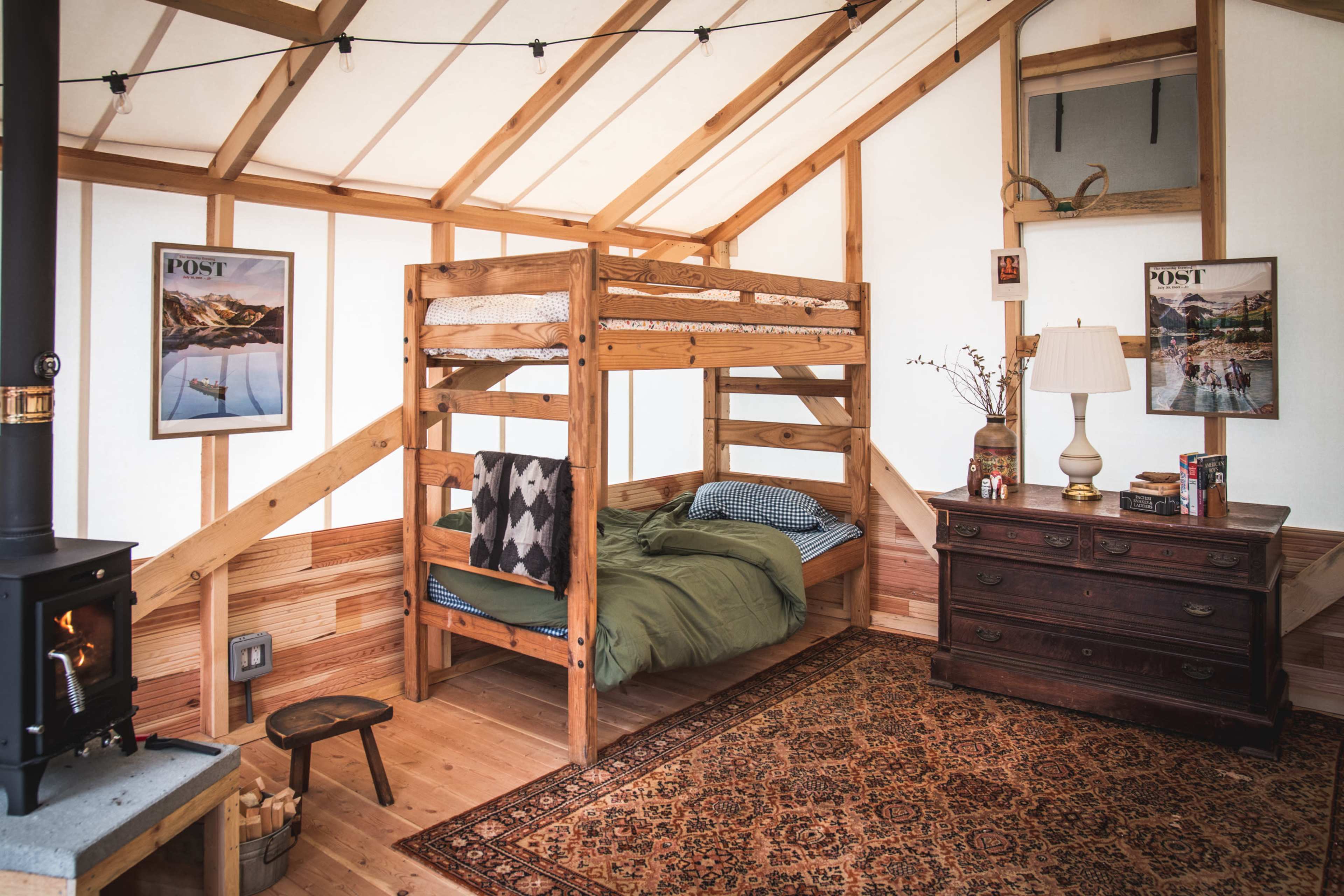 A wooden bunk bed in a cozy room with rustic decor, a vintage dresser, and a small wood stove.