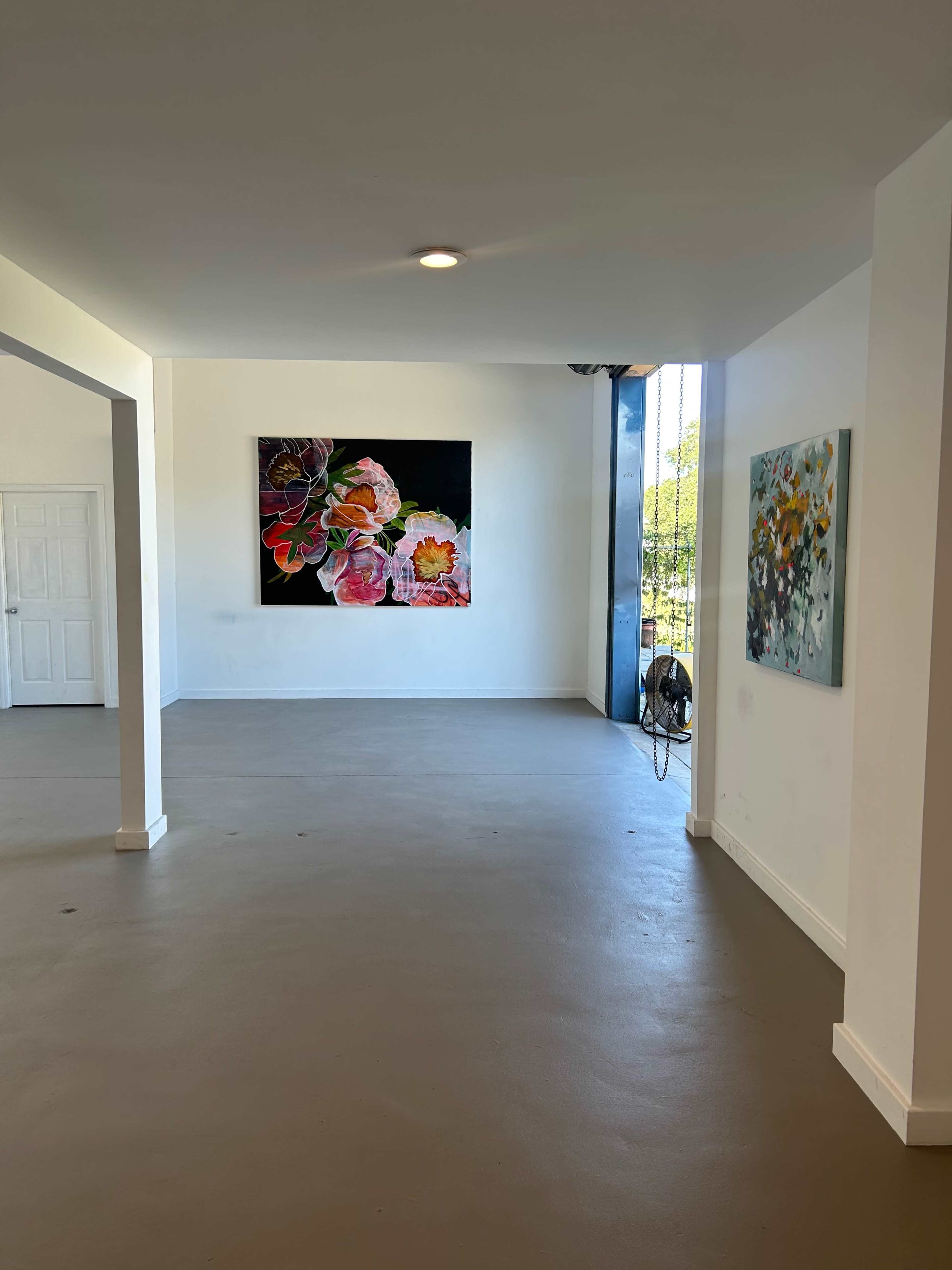 The image shows a spacious interior with light-colored walls, featuring two paintings on the walls and a large window allowing natural light to enter.