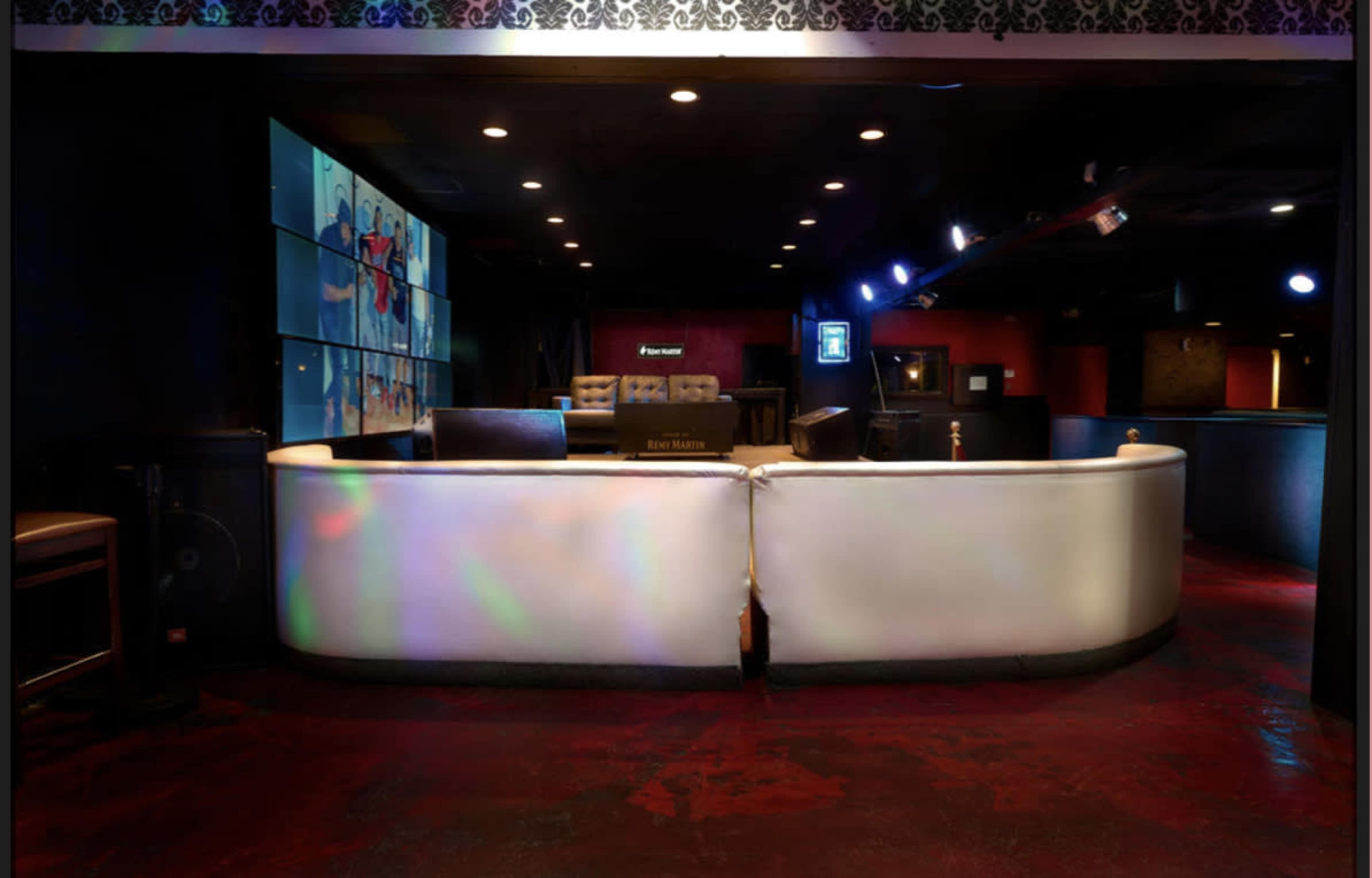 The image shows a modern bar counter in a dimly lit lounge setting, with large screens displaying images on the wall behind it.