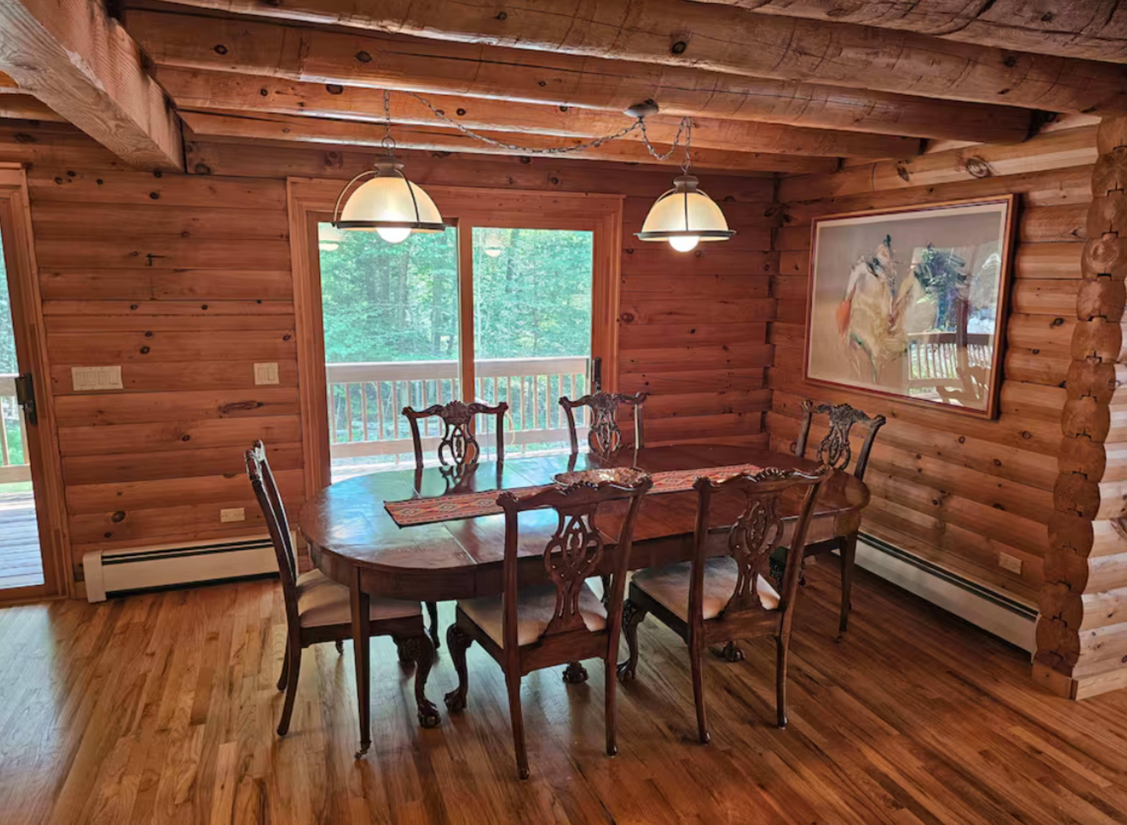 A wooden dining room features a large table surrounded by six chairs, with natural light streaming through two large windows.