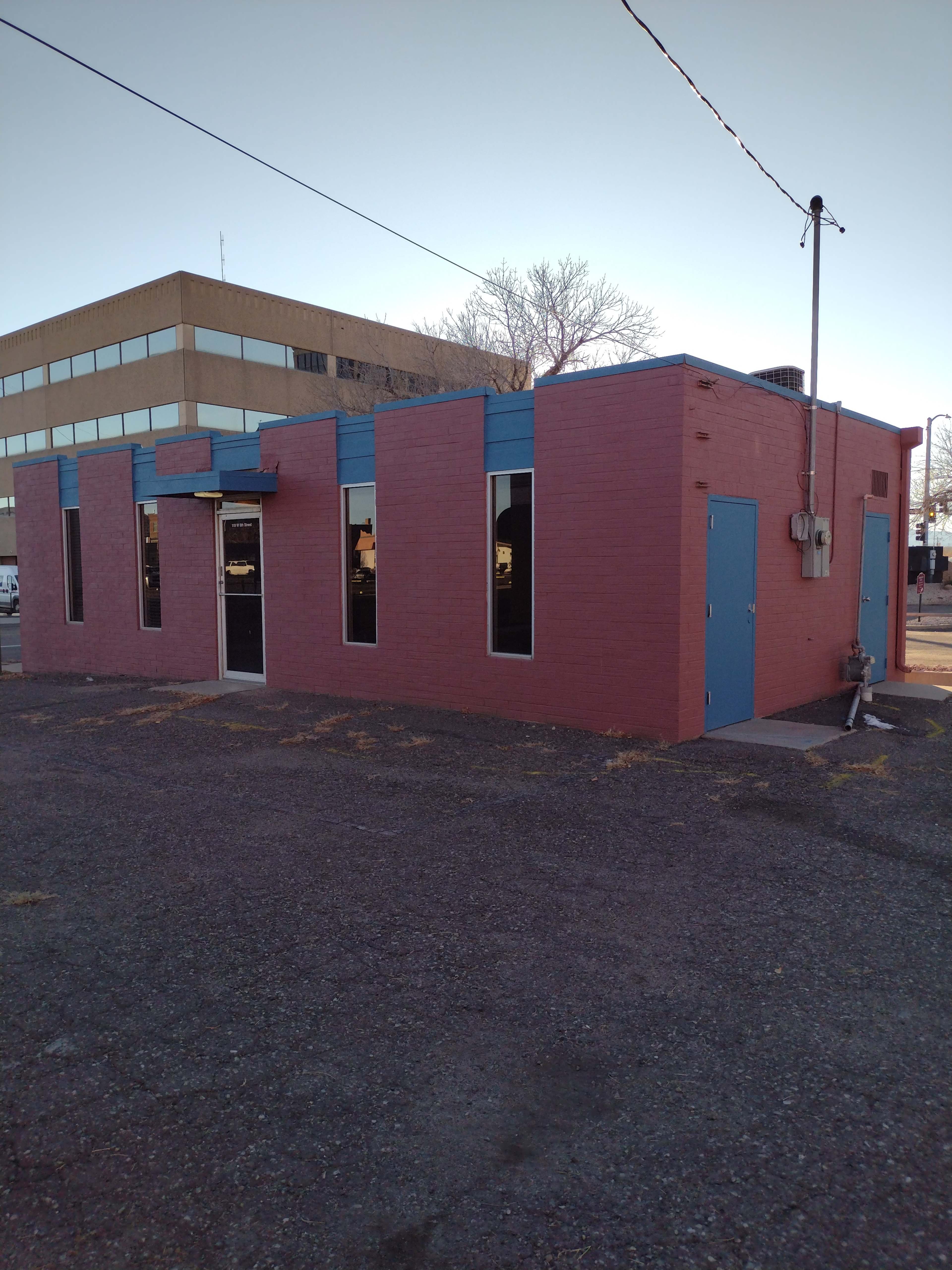 The image shows a single-story building painted in pink with blue accents, featuring multiple windows and a door, situated on a gravel lot next to a taller brick building.