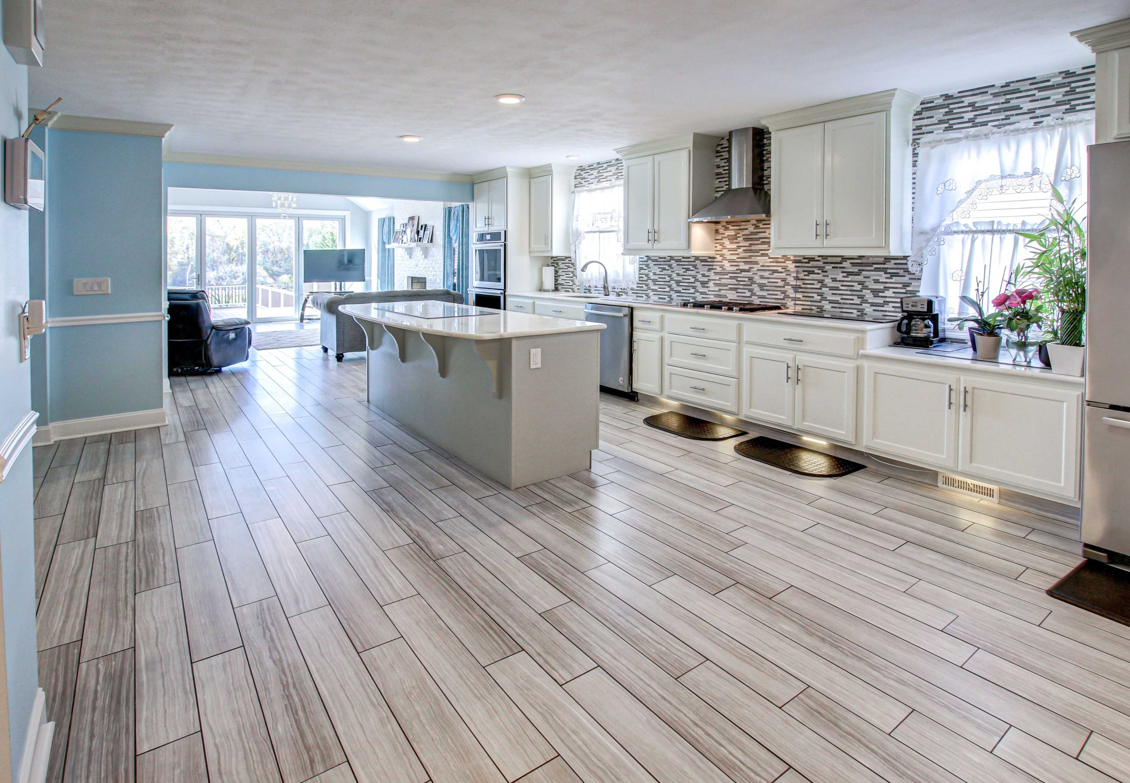 A modern kitchen with light-colored cabinets, a large island, and a tile floor, leading to a well-lit dining area through open doors.