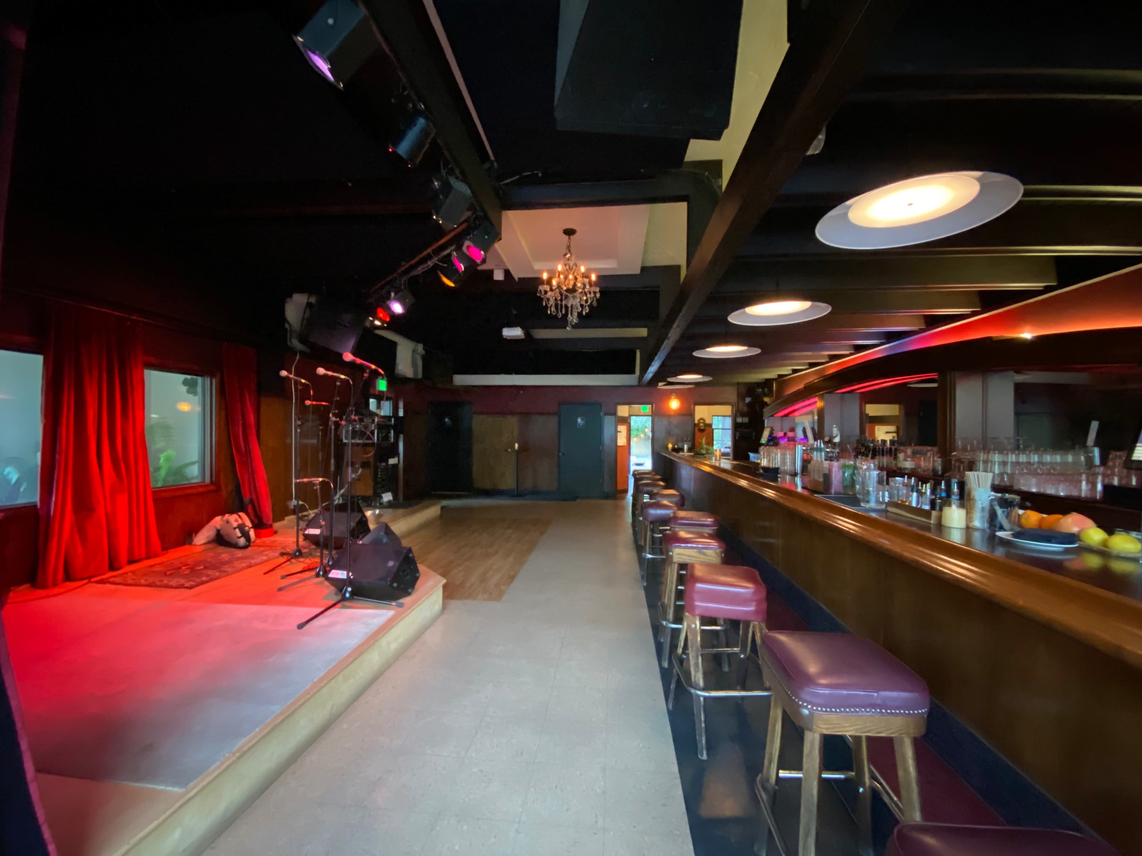 A well-lit interior of a music venue features a stage with red curtains, a bar with stools, and decorative lighting.