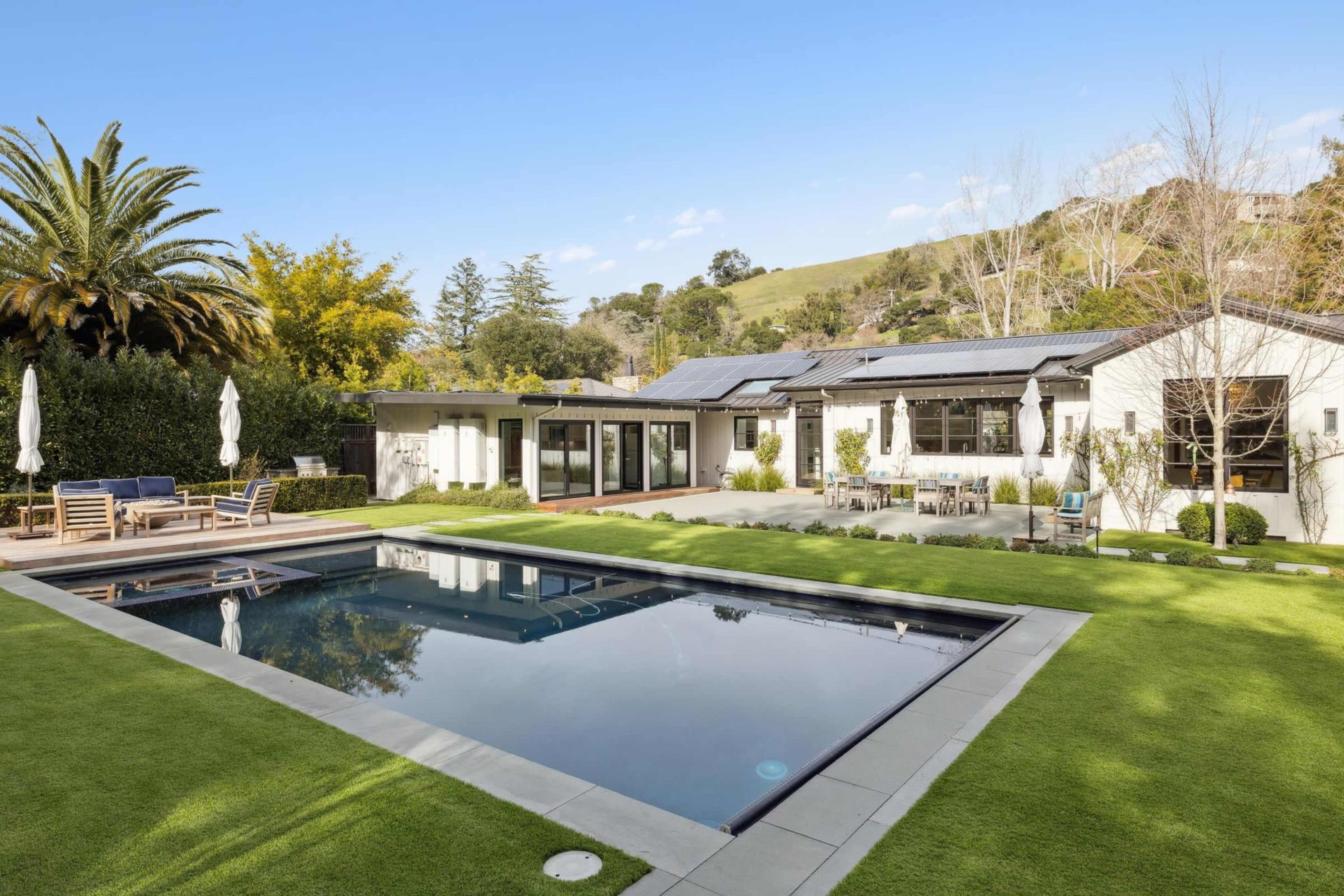 The image shows a modern house with a swimming pool, surrounded by well-maintained lawns and palm trees in a sunny setting.