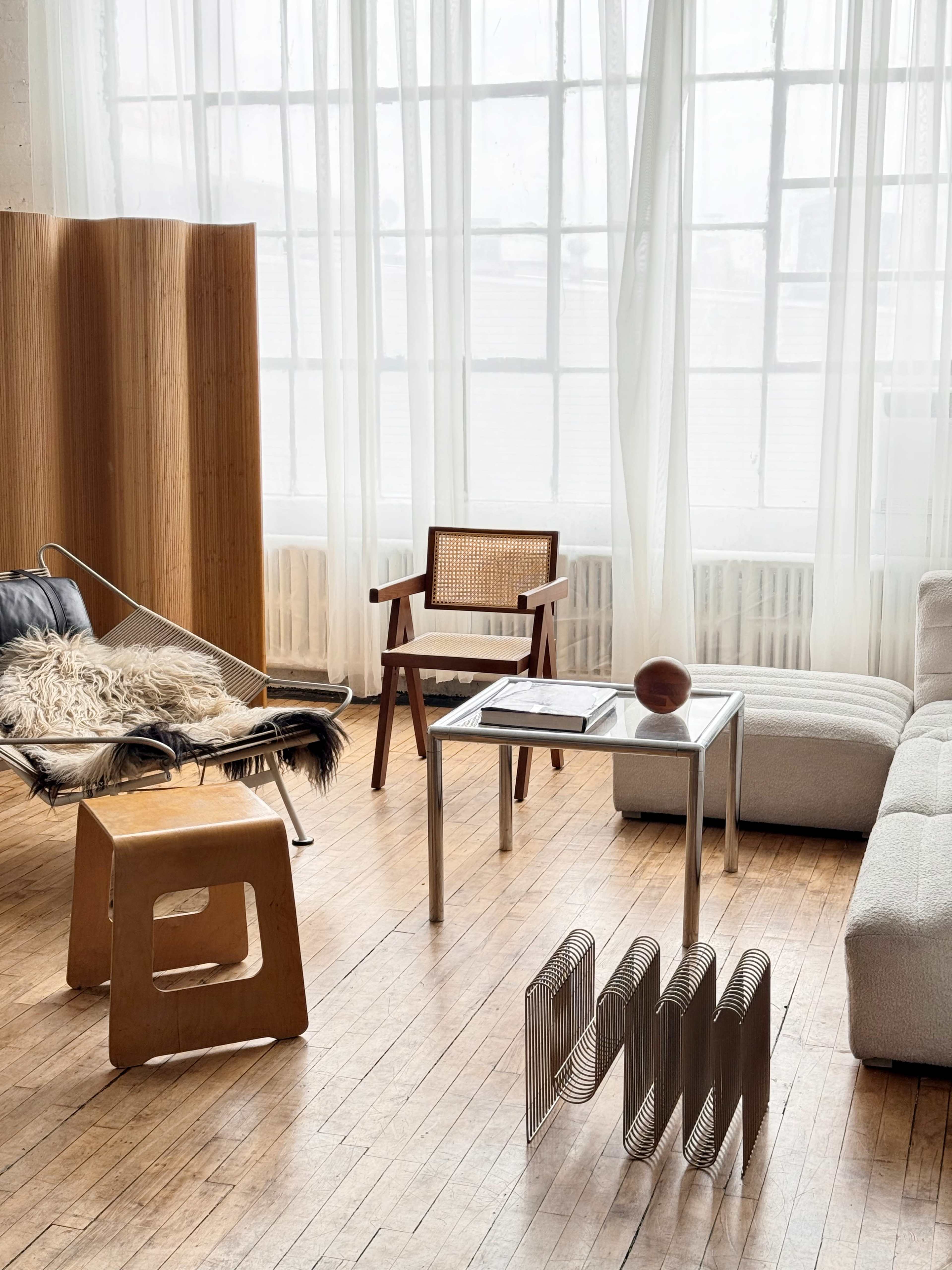 The room features a light-filled space with wooden flooring, showcasing a mix of modern furniture pieces, including a sleek gray couch, a wooden chair, a minimalist table, and a unique wooden stool.
