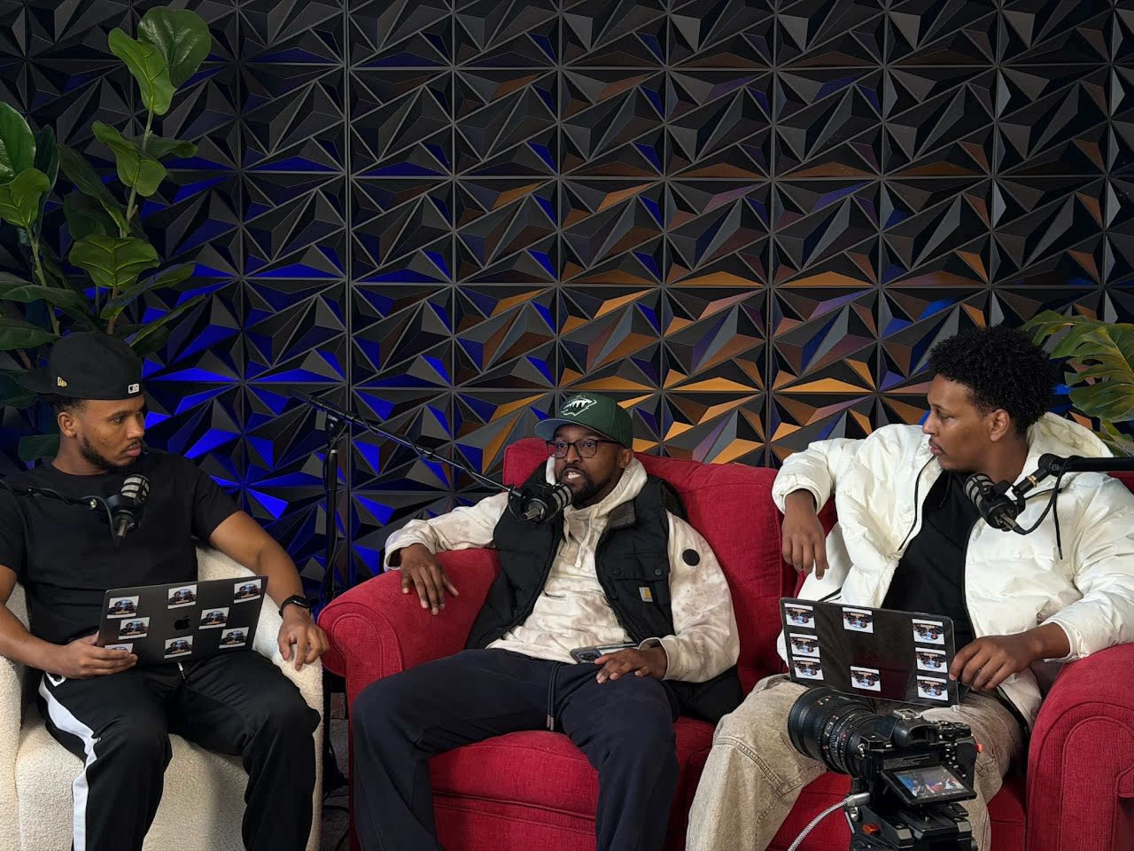 Three men are seated on red couches in a studio setting, engaged in a discussion while holding notebooks and microphones.