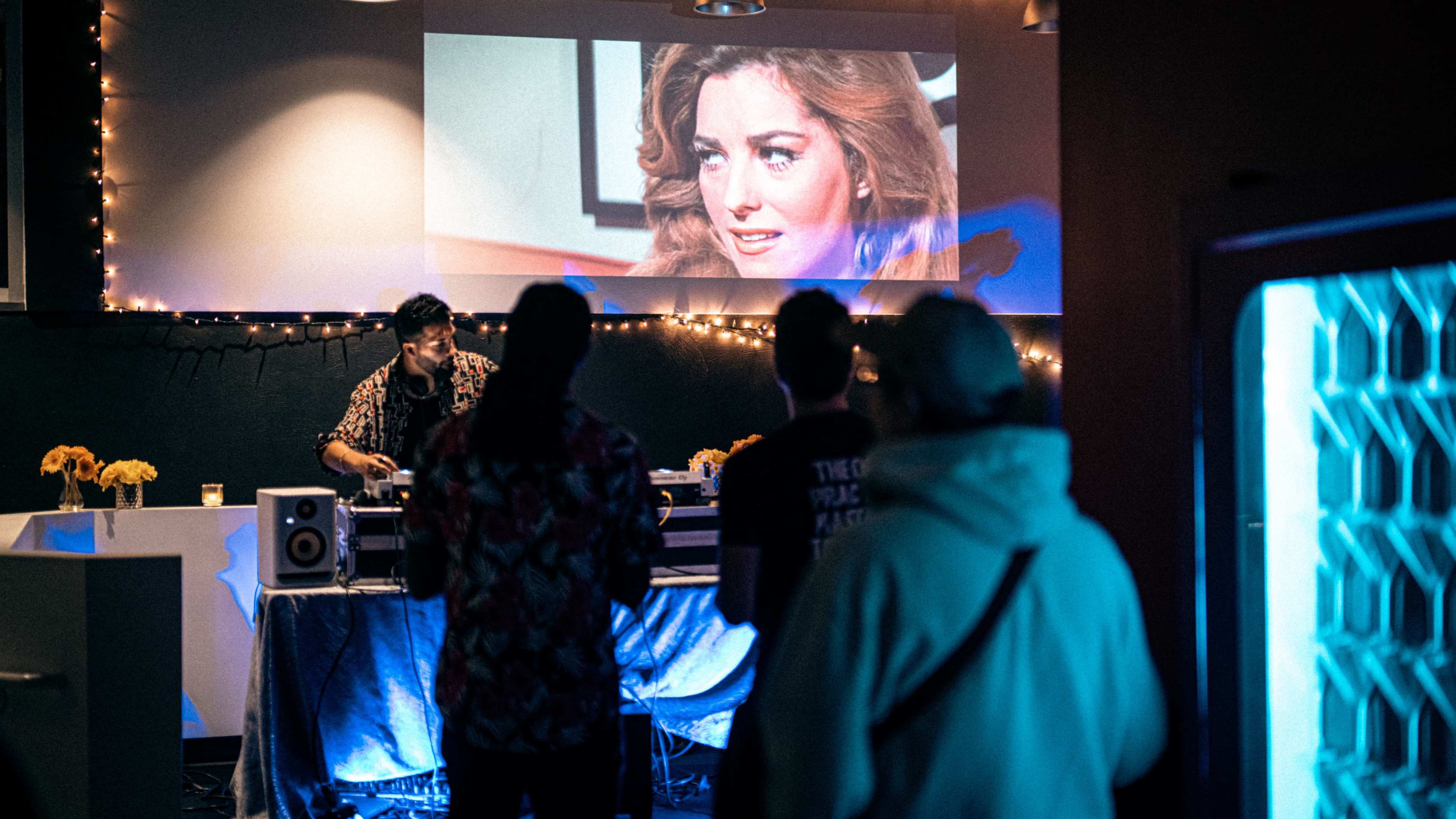 A DJ performs at a small event while an old movie scene plays on a screen behind them, with attendees watching and socializing.