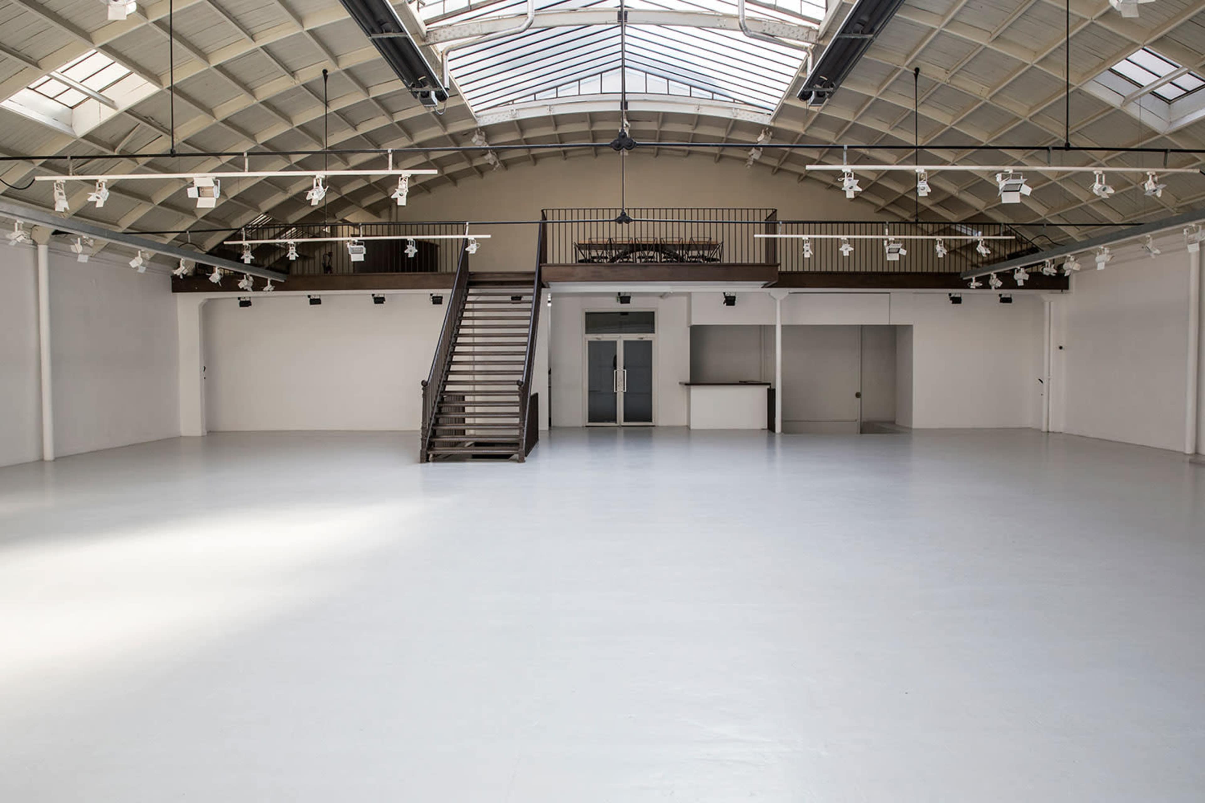 An empty, spacious interior with a high ceiling, large windows, and a staircase leading to a balcony.