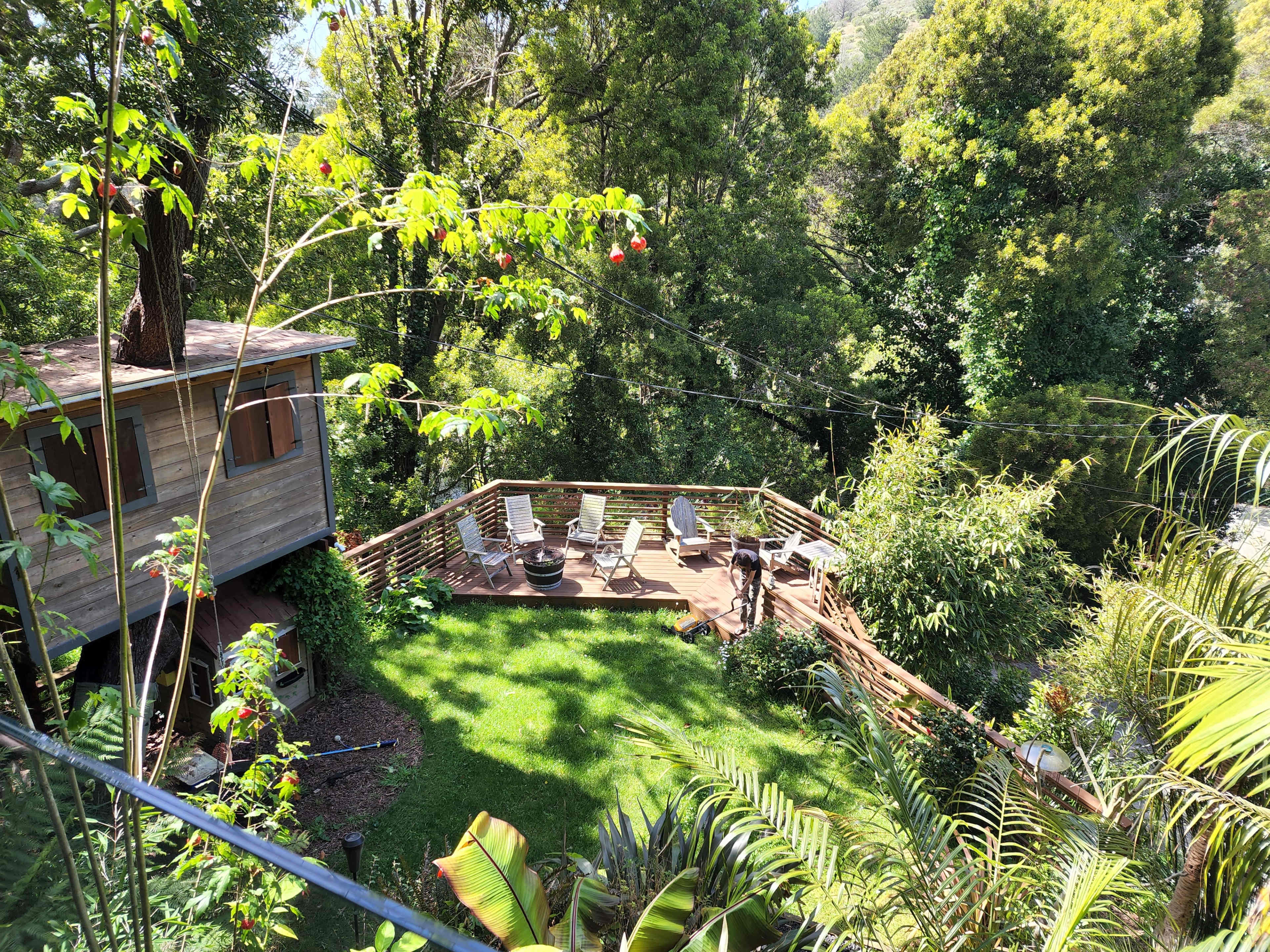 The image shows a wooden deck surrounded by greenery, with chairs arranged for seating and a treehouse nearby.