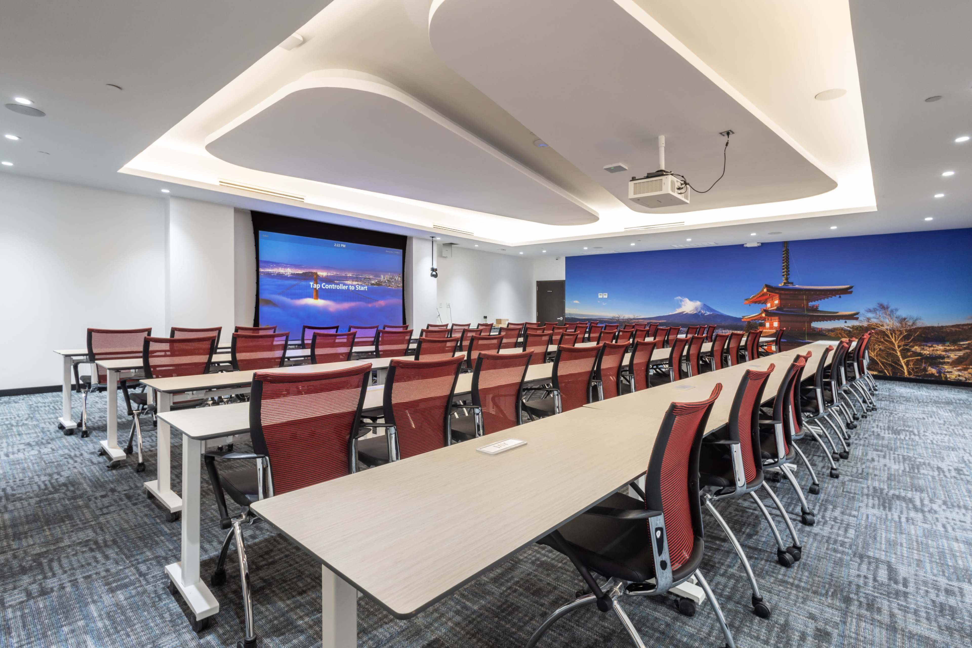 A modern conference room with rows of red chairs and long tables, featuring a large screen at one end and a scenic mural on the wall.
