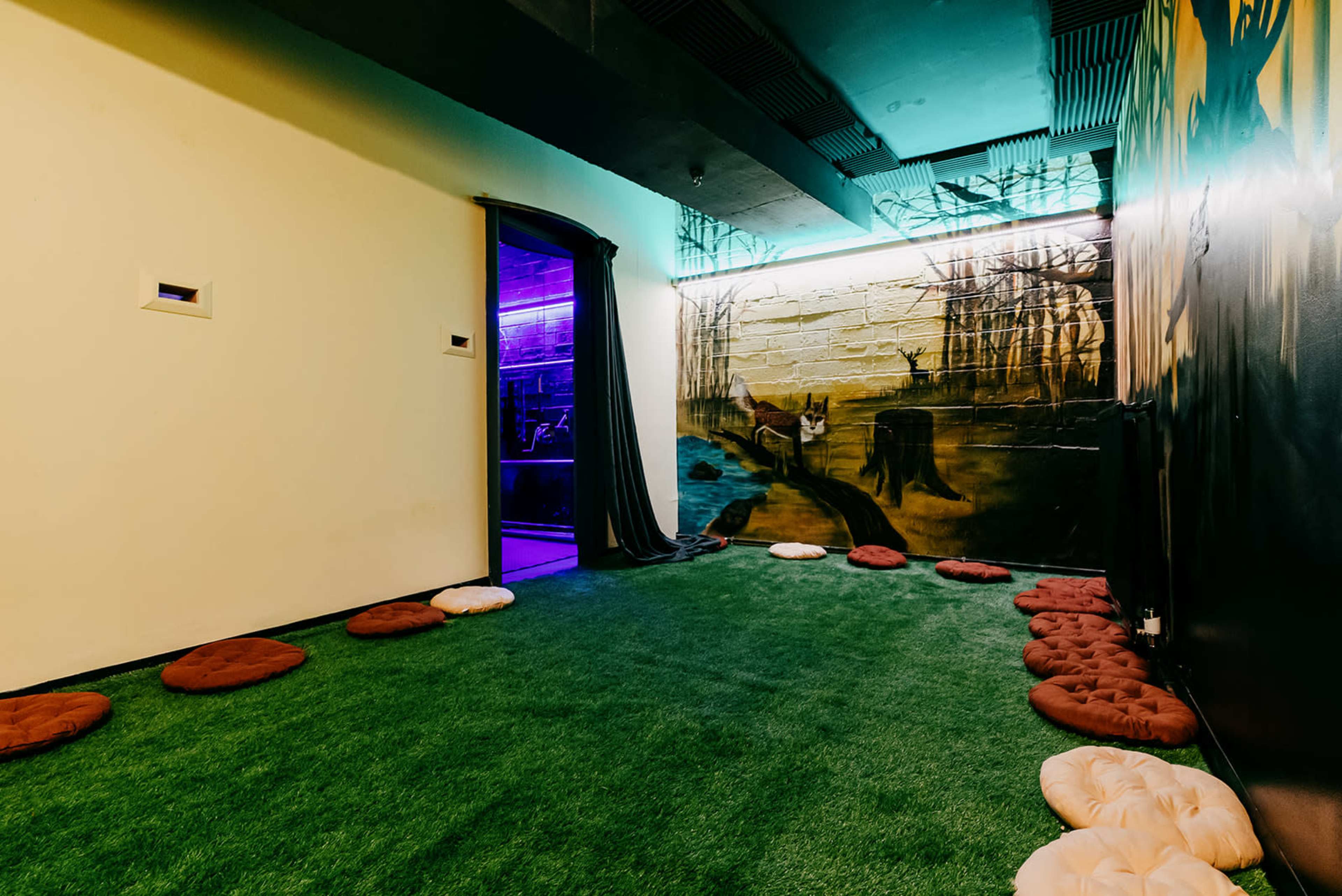 A small room with artificial grass flooring, scattered cushions, and a mural of a natural scene on one wall.