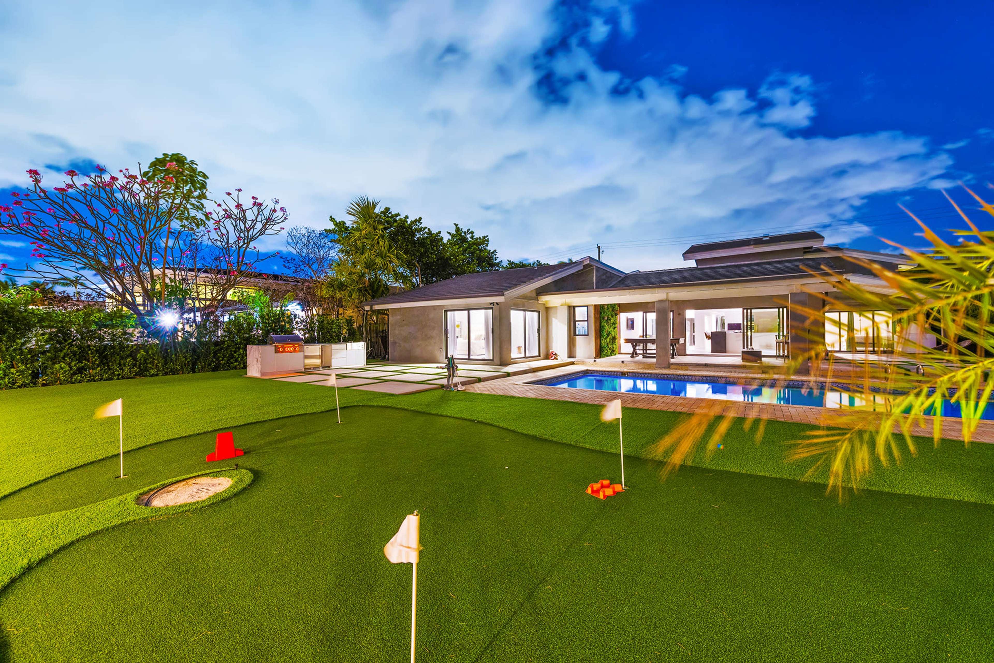 A modern house with a swimming pool and a green, manicured lawn featuring putting greens and landscaping under a blue sky.