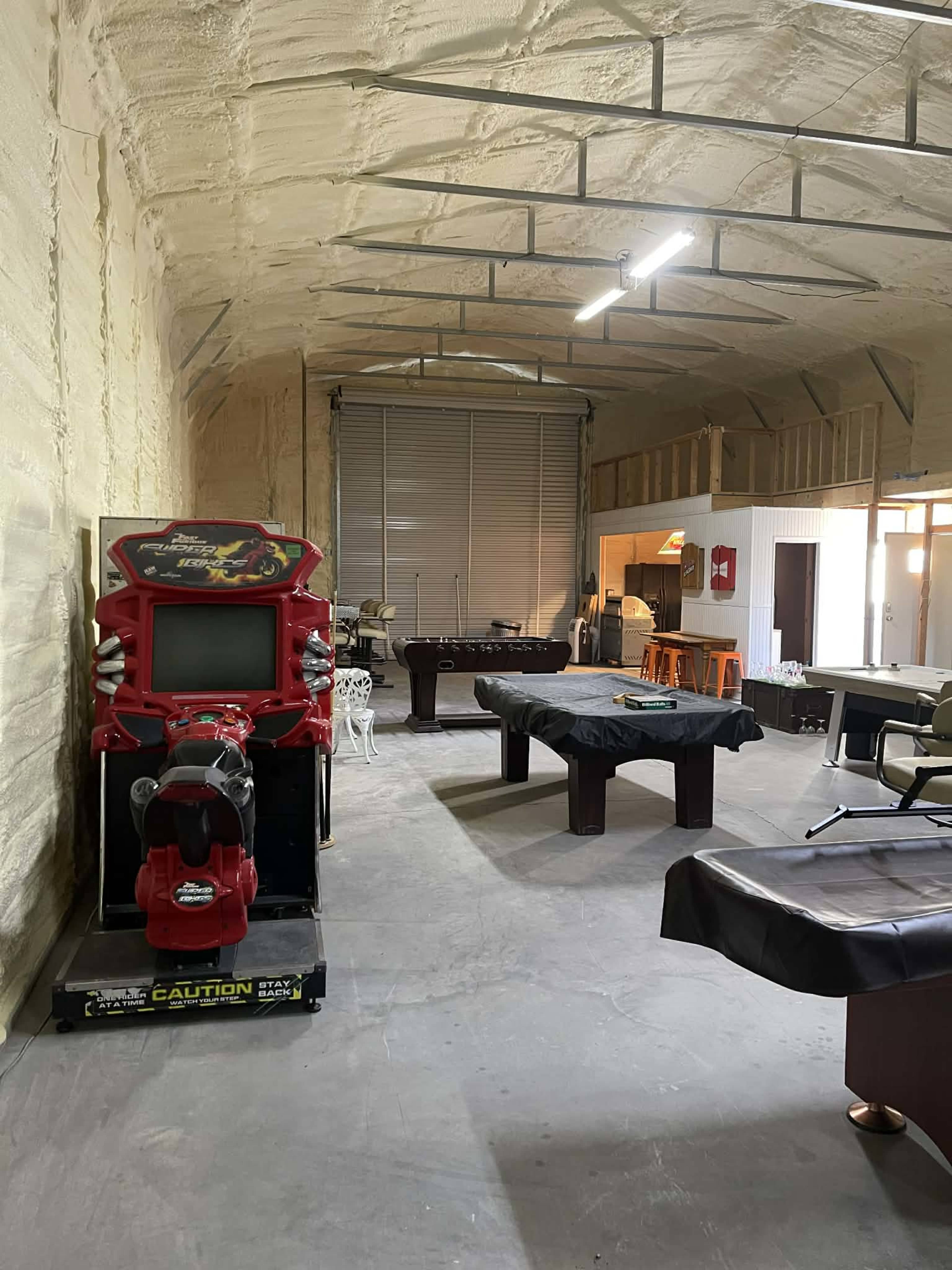 The image shows a spacious indoor area with a red arcade machine, a pool table covered with a black cloth, and various chairs and tables arranged around the room.