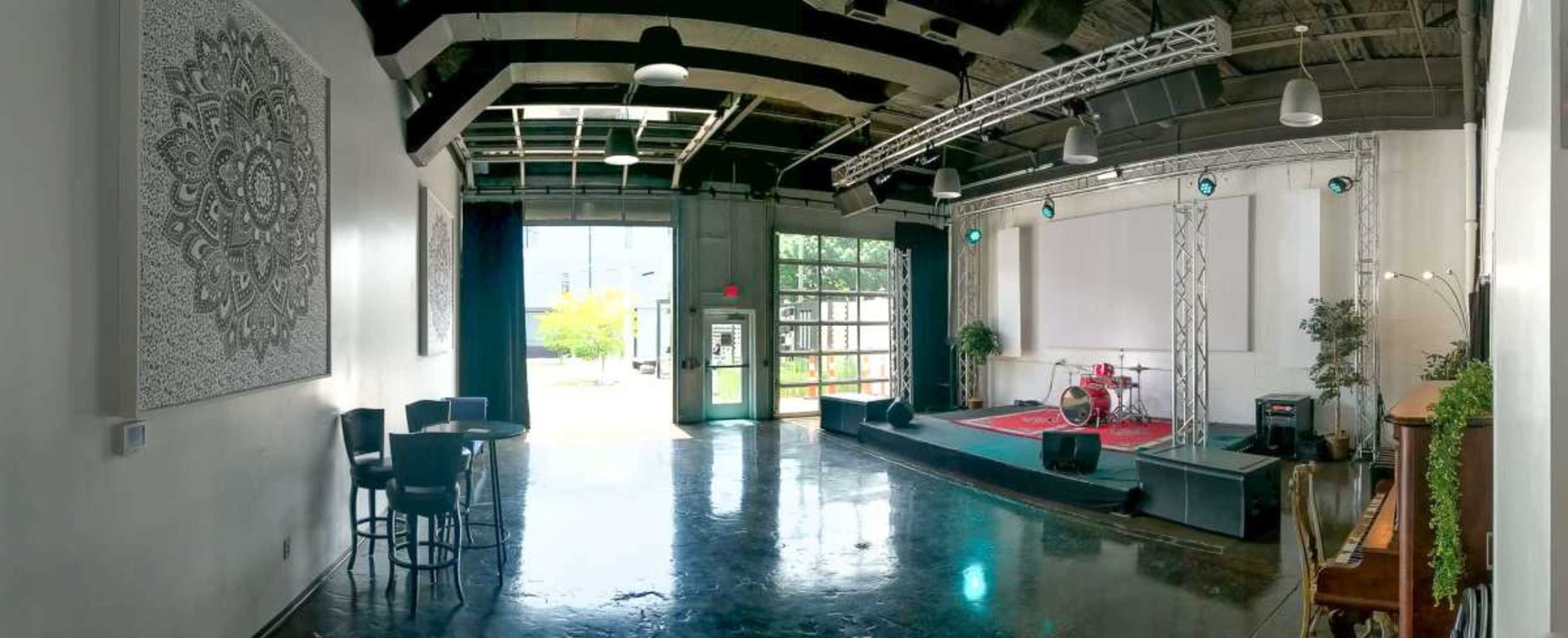 The image shows a spacious performance venue with a stage featuring musical equipment, large windows at the entrance, and decorative elements on the walls.