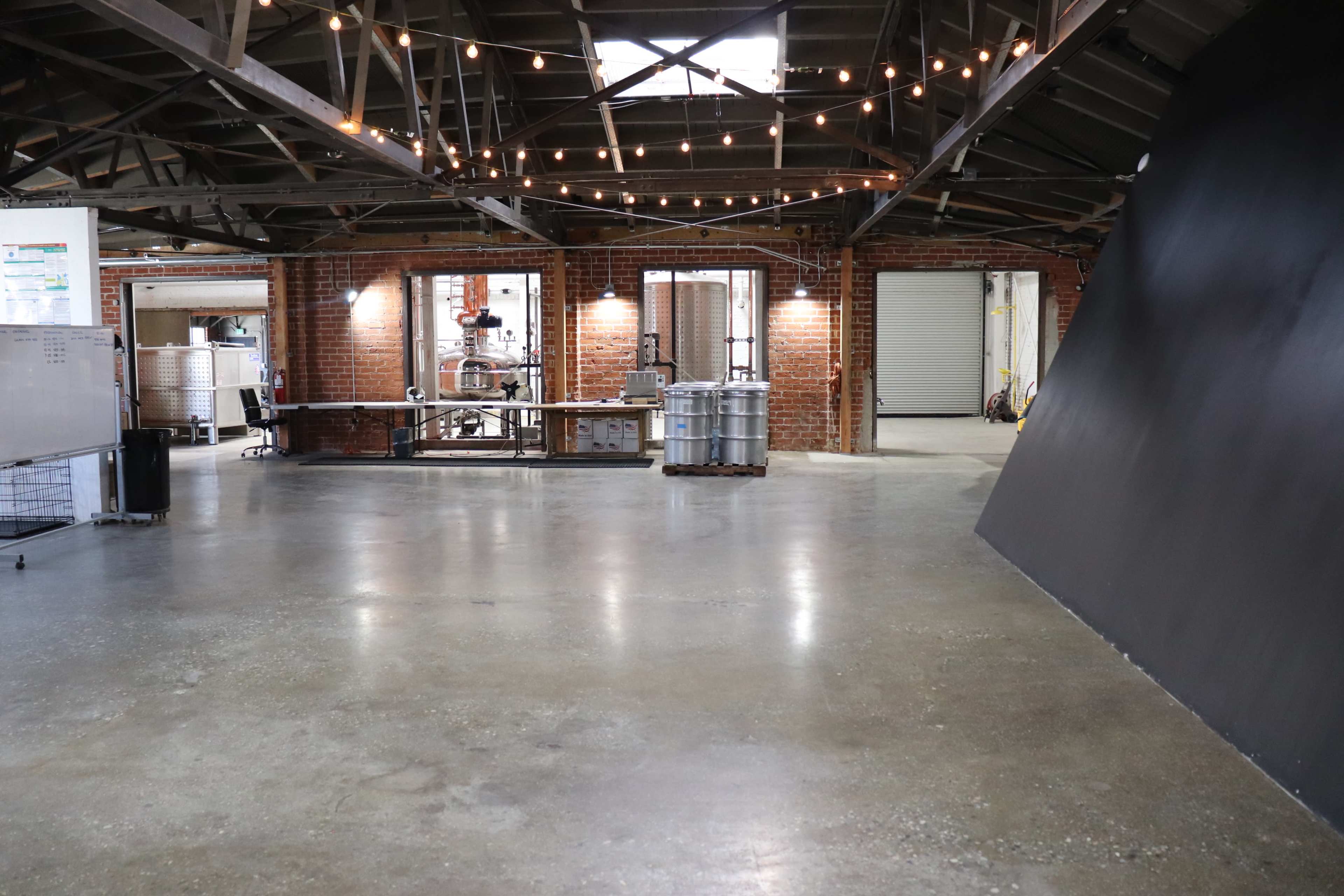 A spacious industrial interior with exposed brick walls, large windows, and overhead string lights, featuring a concrete floor and stainless steel work tables.
