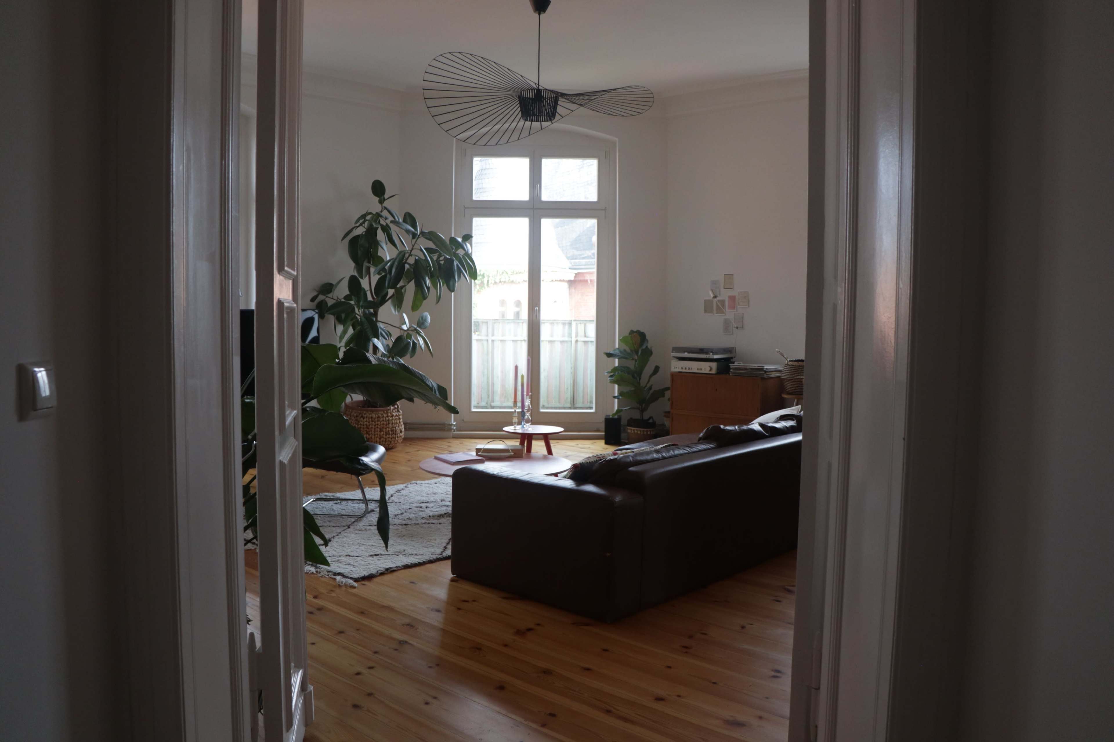 A view of a living room with a brown sofa, a large plant, and a light fixture, seen through an open doorway.