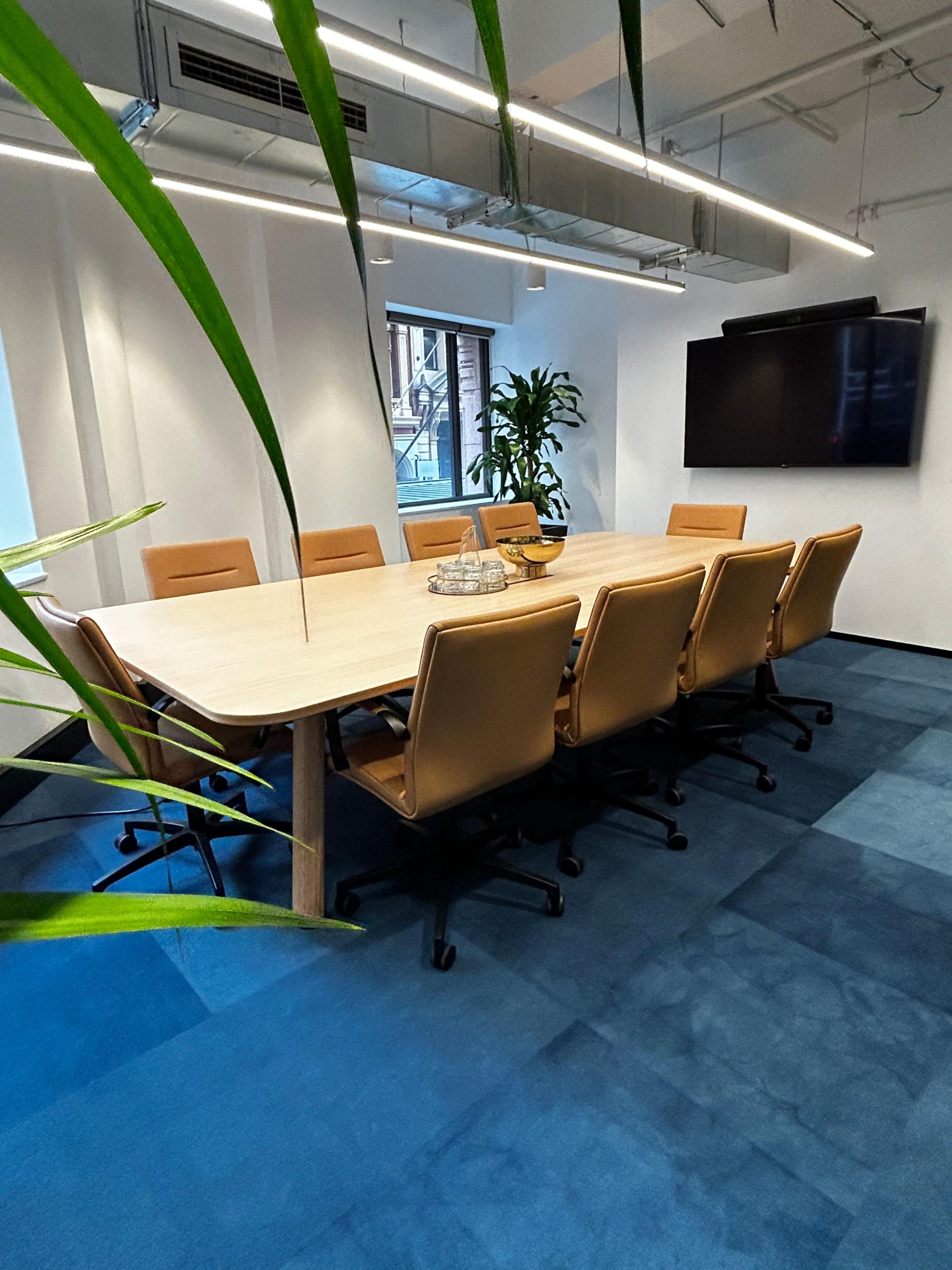 A spacious conference room features a large wooden table surrounded by ergonomic chairs, with plants and a wall-mounted screen in the background.