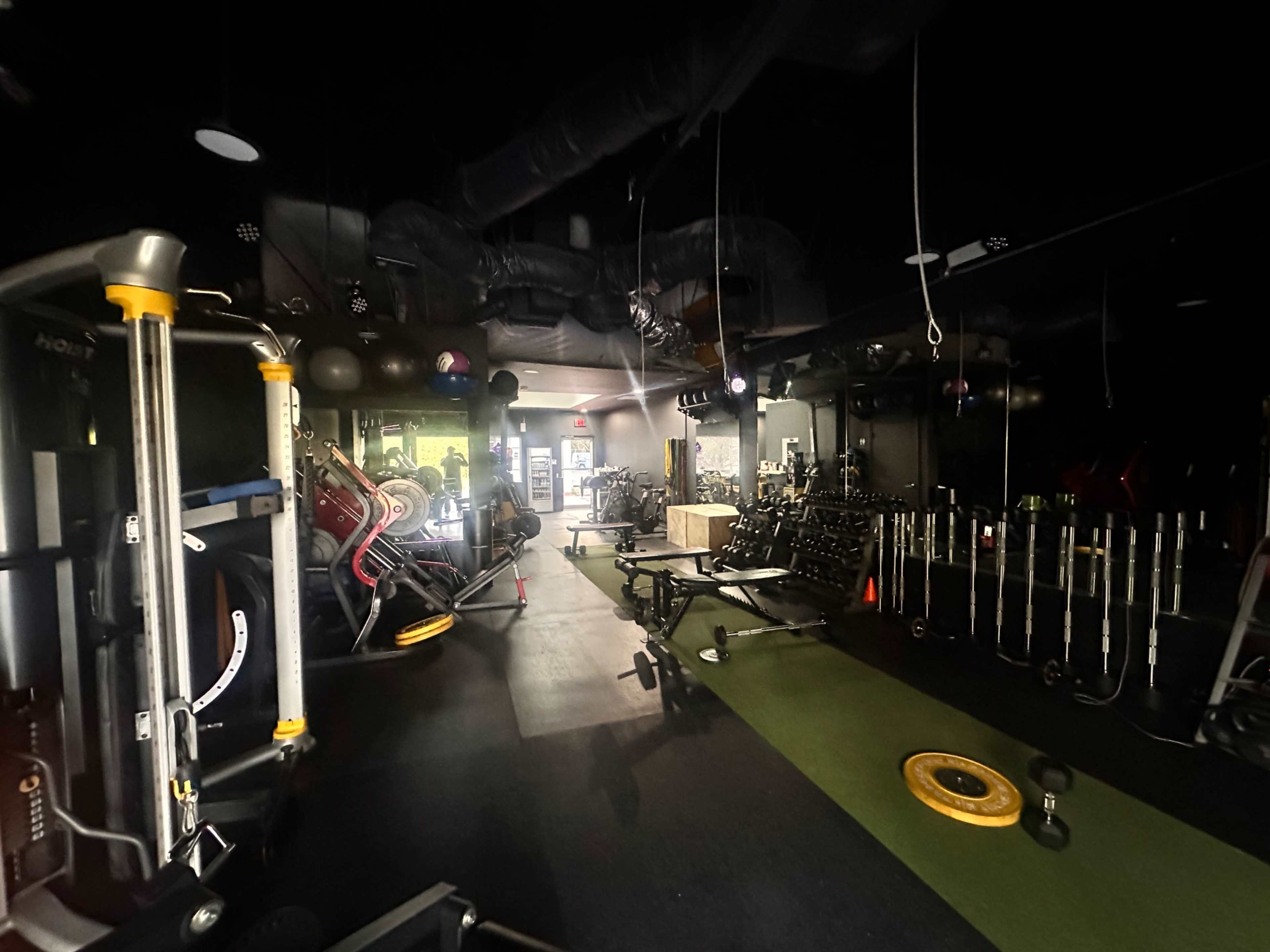 The image shows a dimly lit gym with various exercise equipment arranged along the walls and a green turf area in the center.