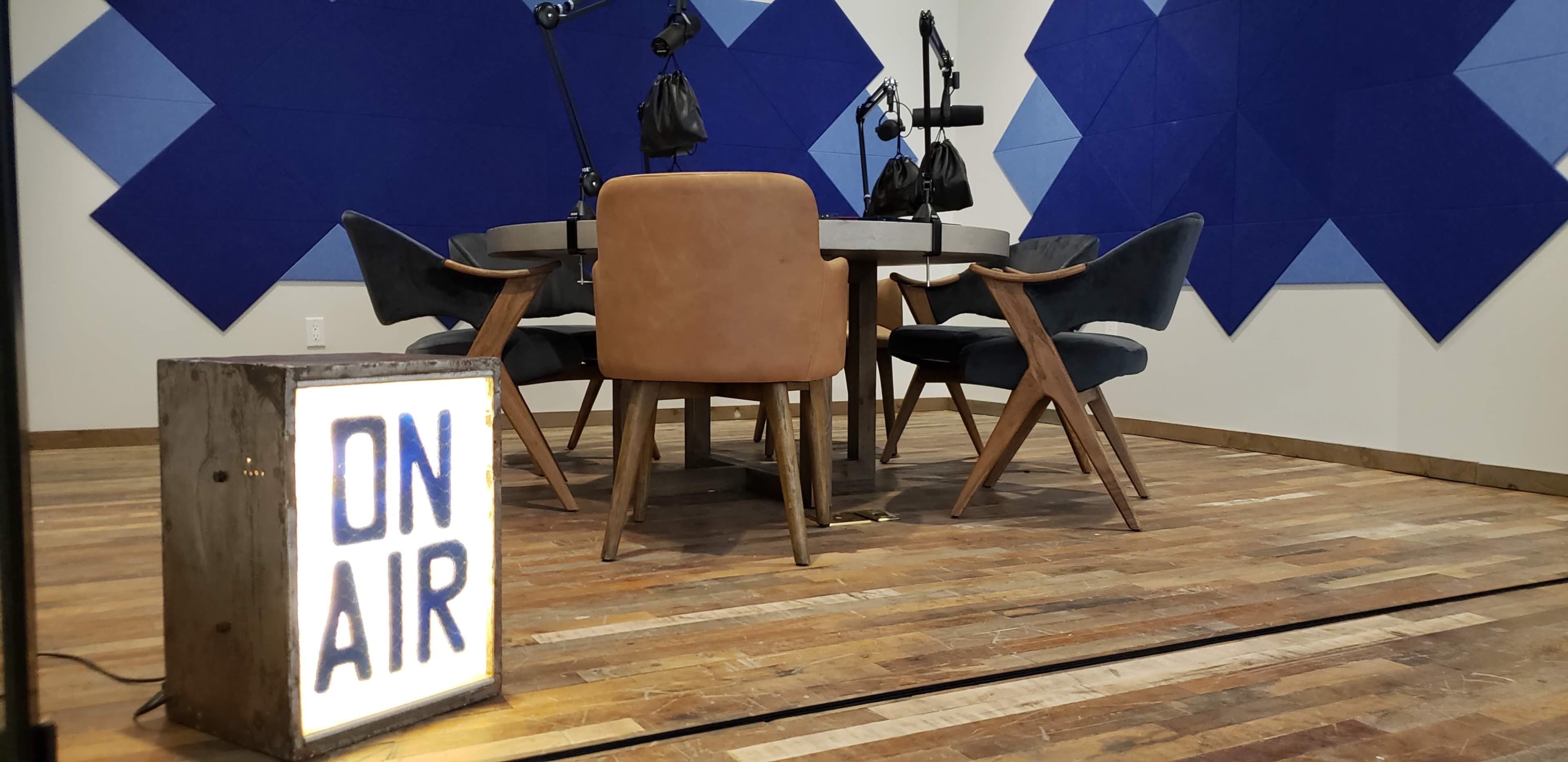 A recording studio features a circular table surrounded by chairs, with a "ON AIR" sign illuminated in the foreground.