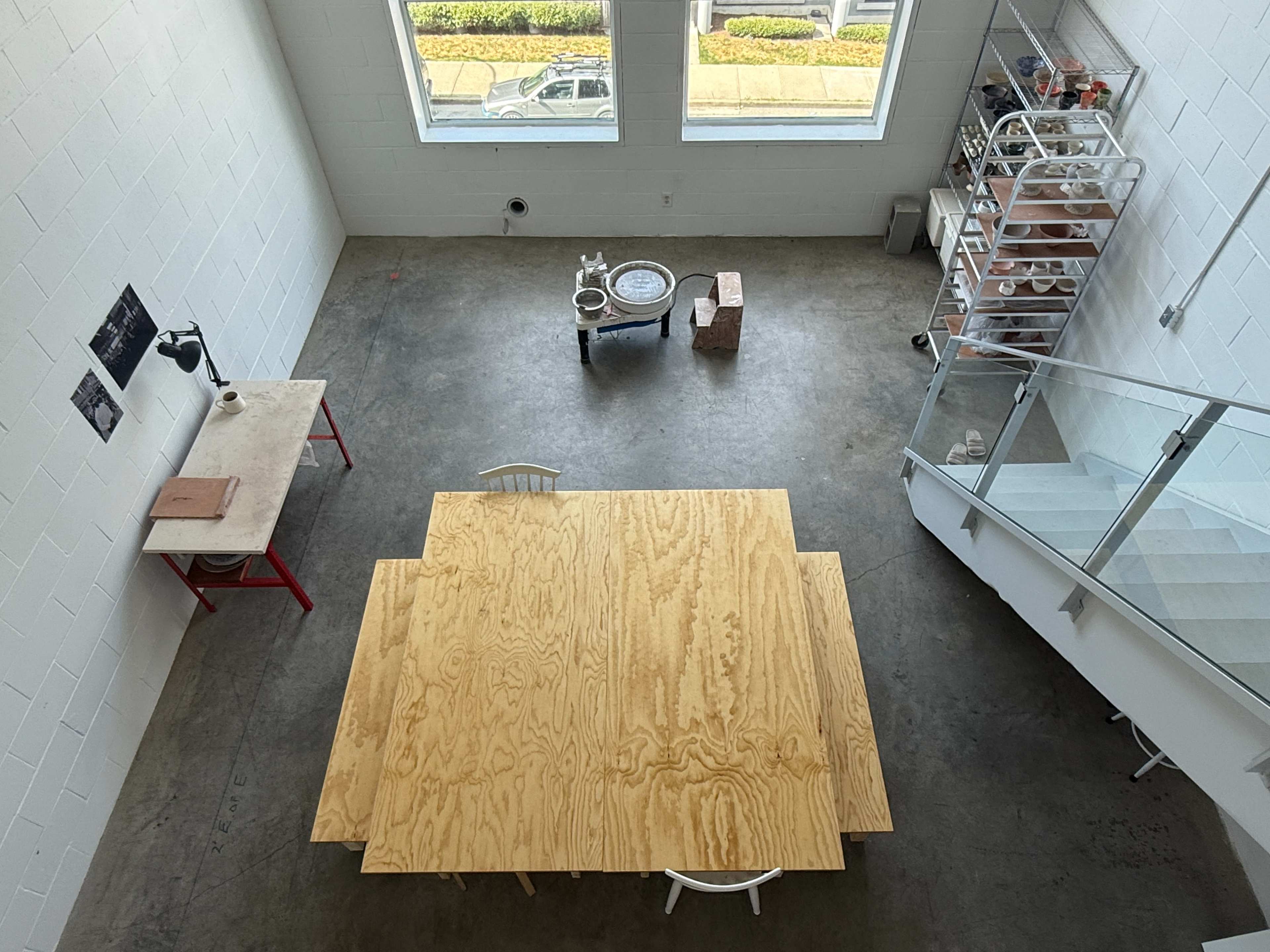 The image shows an open, minimalist studio space with a large wooden table, a lamp, and various items placed on shelves and a small cart.