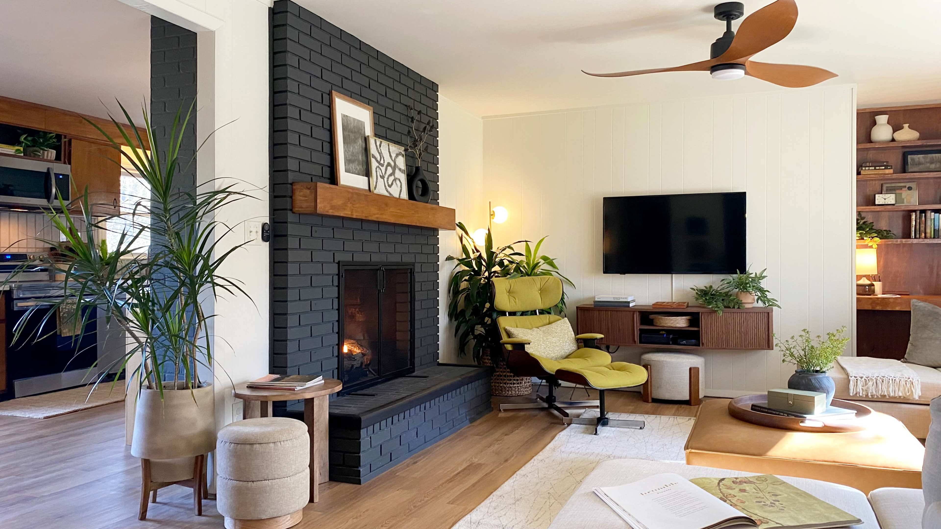 The living room features a dark brick fireplace, a mounted television, a green accent chair, and various indoor plants.