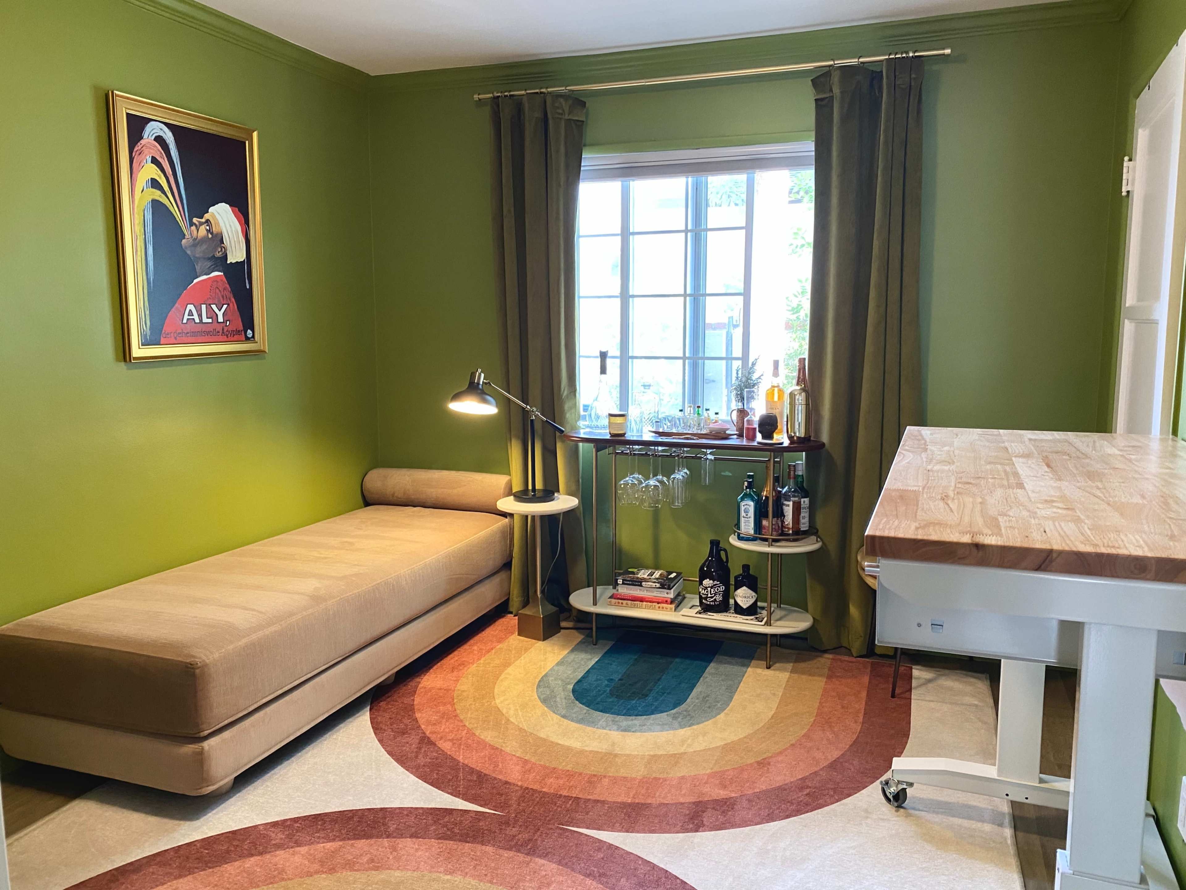 A cozy room with green walls, featuring a sofa, a bar cart with bottles, and a window illuminated by natural light.