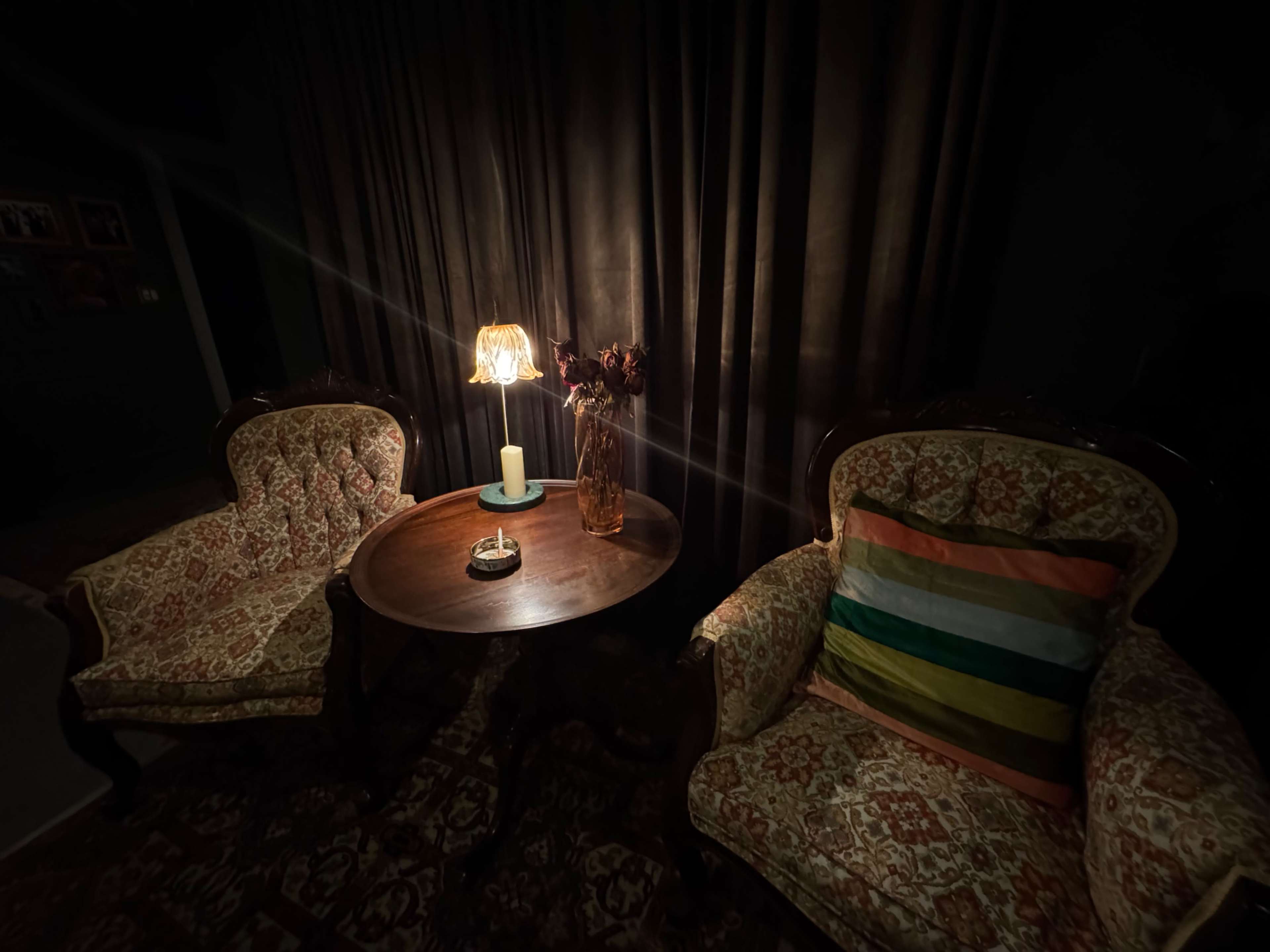 The image shows a dimly lit room with two ornate chairs, a round table, a lit lamp, and a vase with dried flowers.