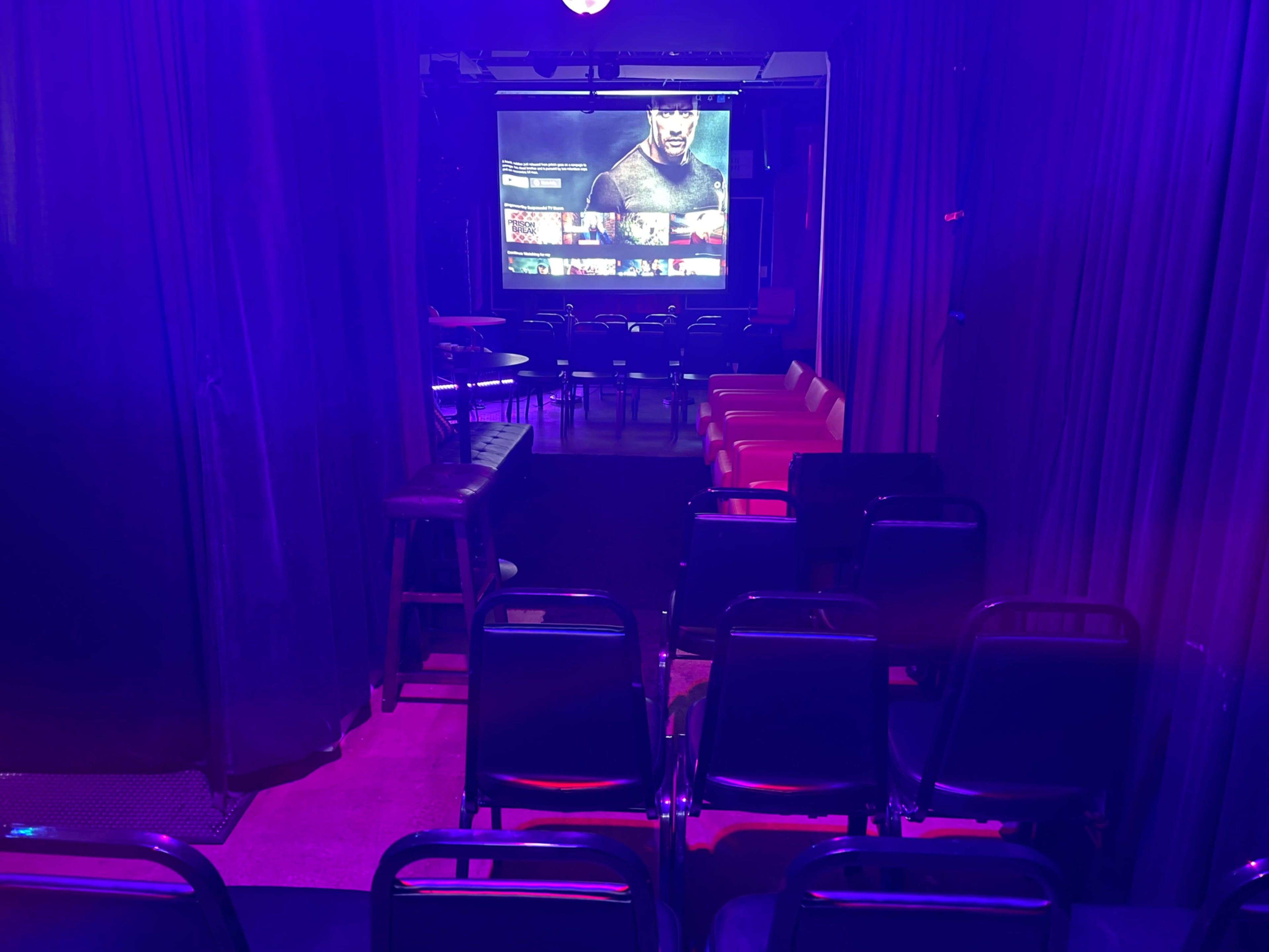 The image shows a dimly lit room with rows of chairs facing a large screen displaying a person, while purple and blue lighting creates a vibrant atmosphere.