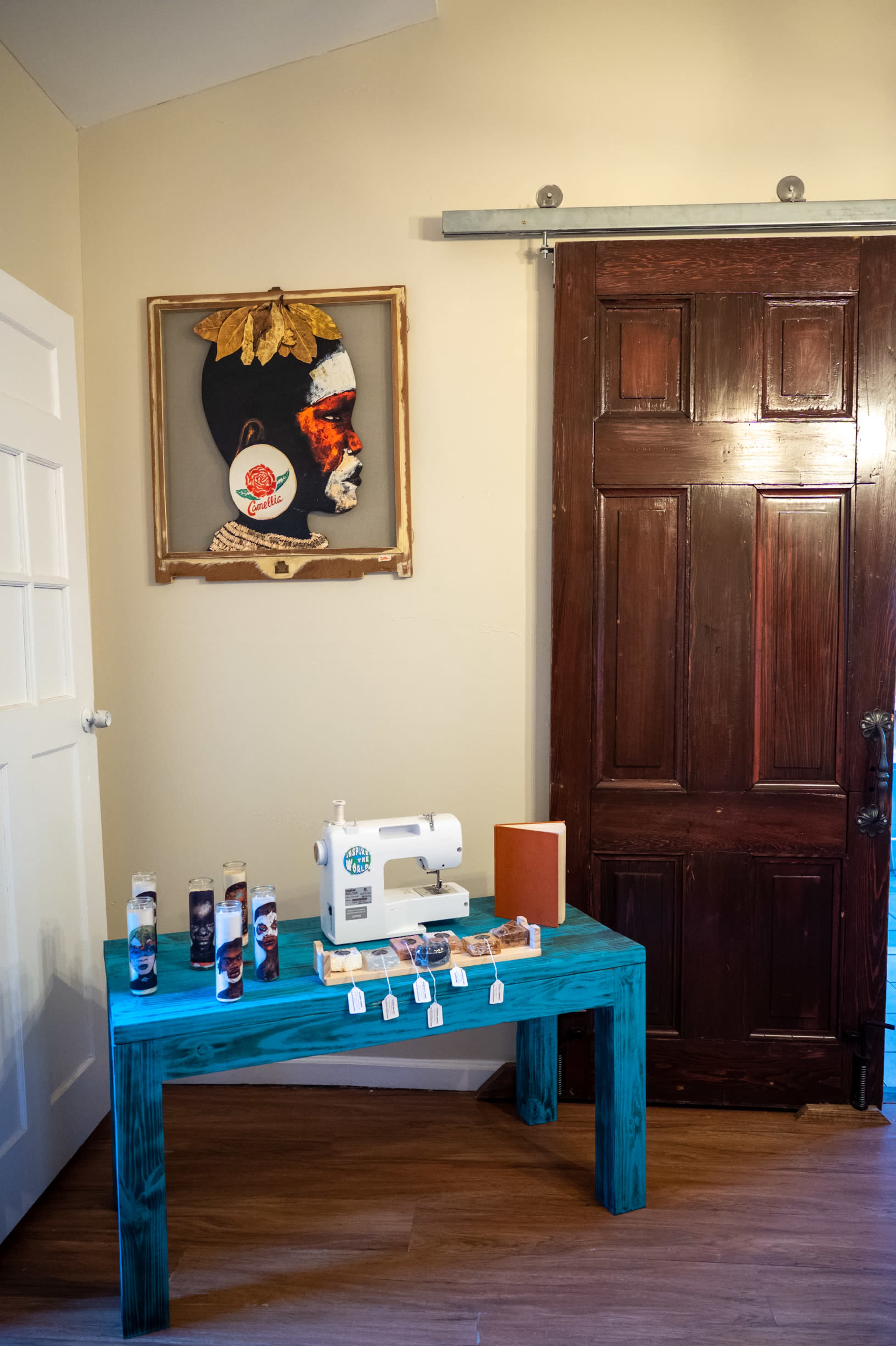 A turquoise table with a sewing machine, a notebook, and decorative glass jars sits beside a wooden door and an art piece featuring a profile portrait.