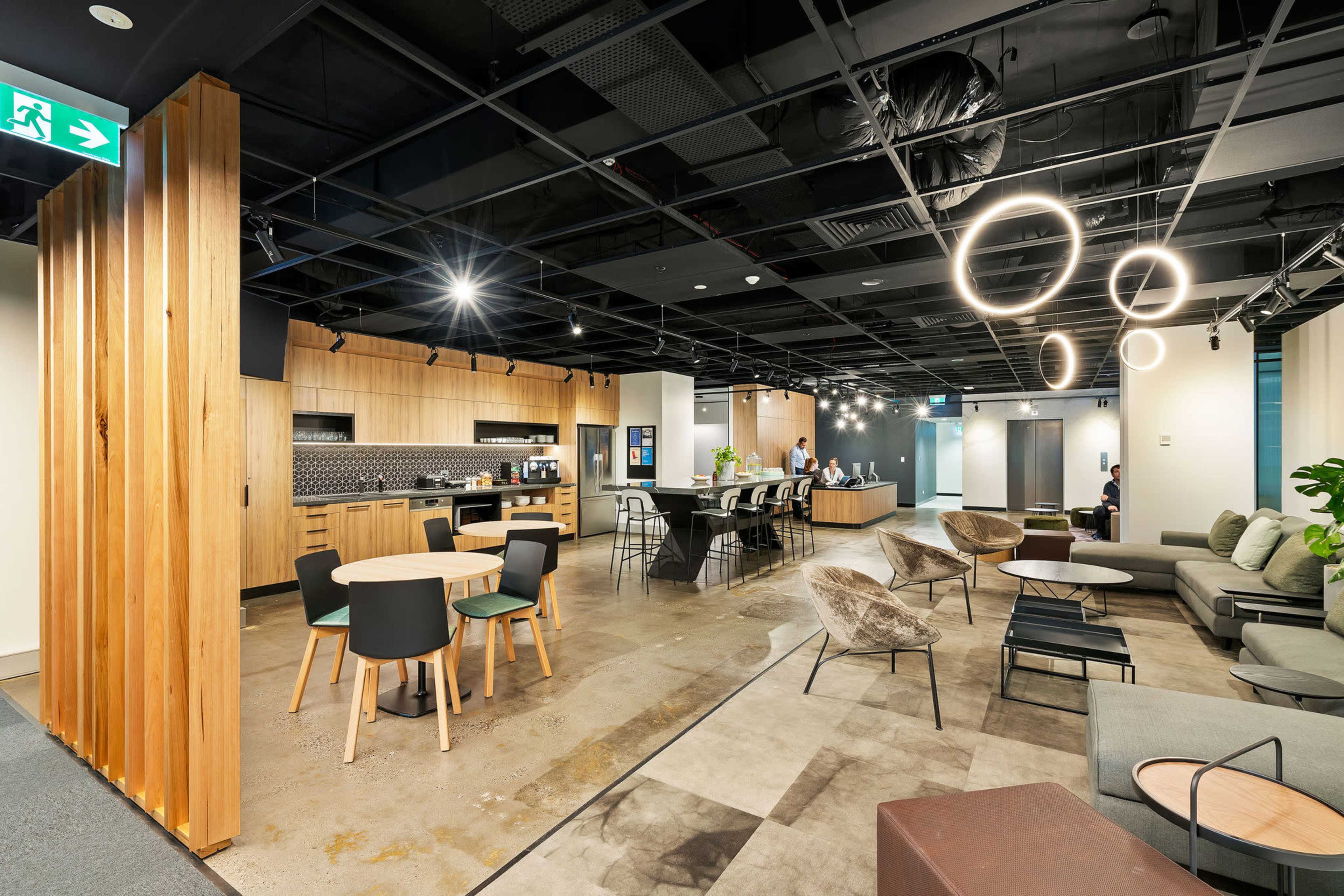 The image shows a modern office space featuring a kitchen area, workstations, and seating arrangements, with wooden and metal furnishings.