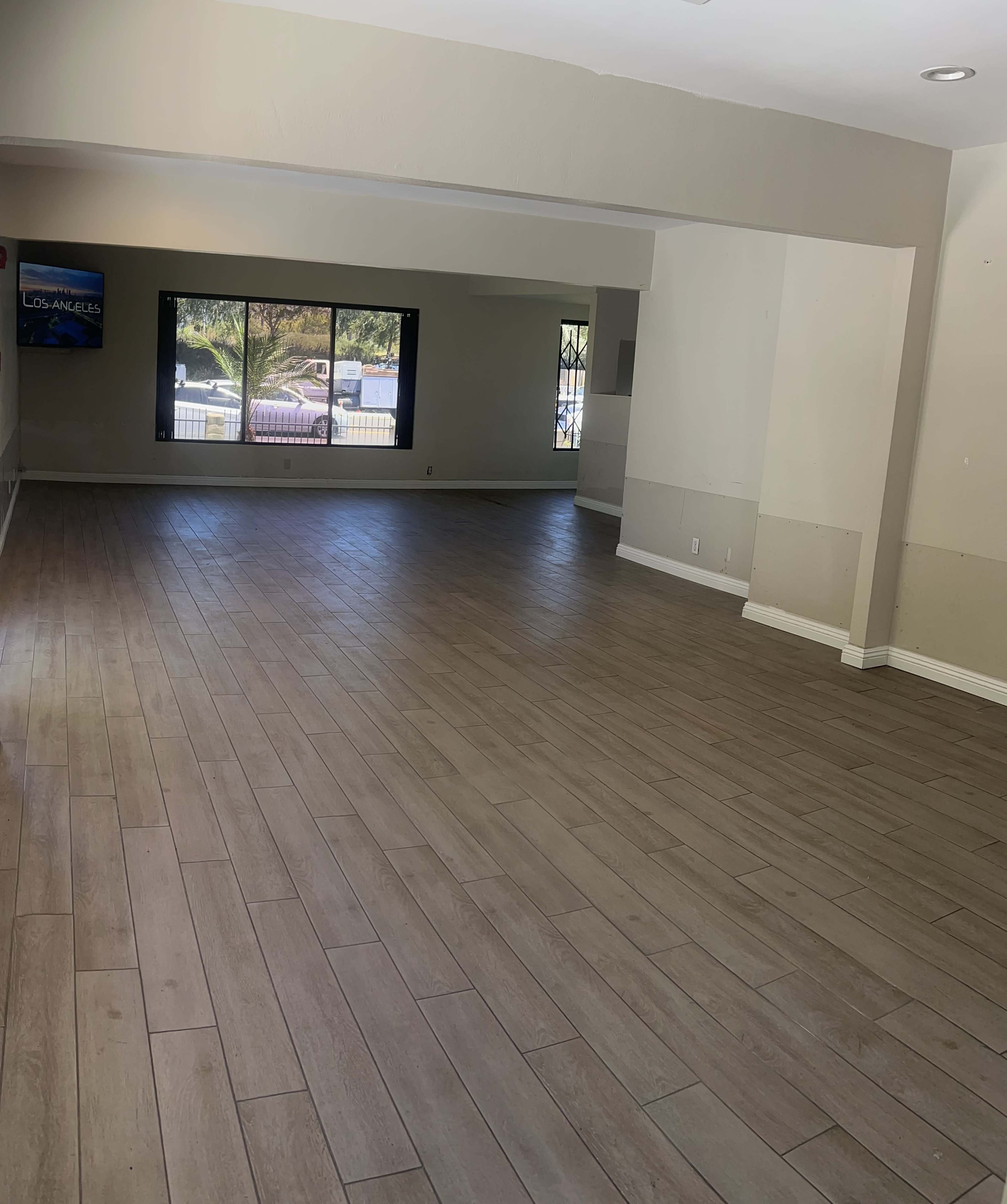 The image shows a spacious room with light-colored wooden flooring and large windows that allow natural light to enter.